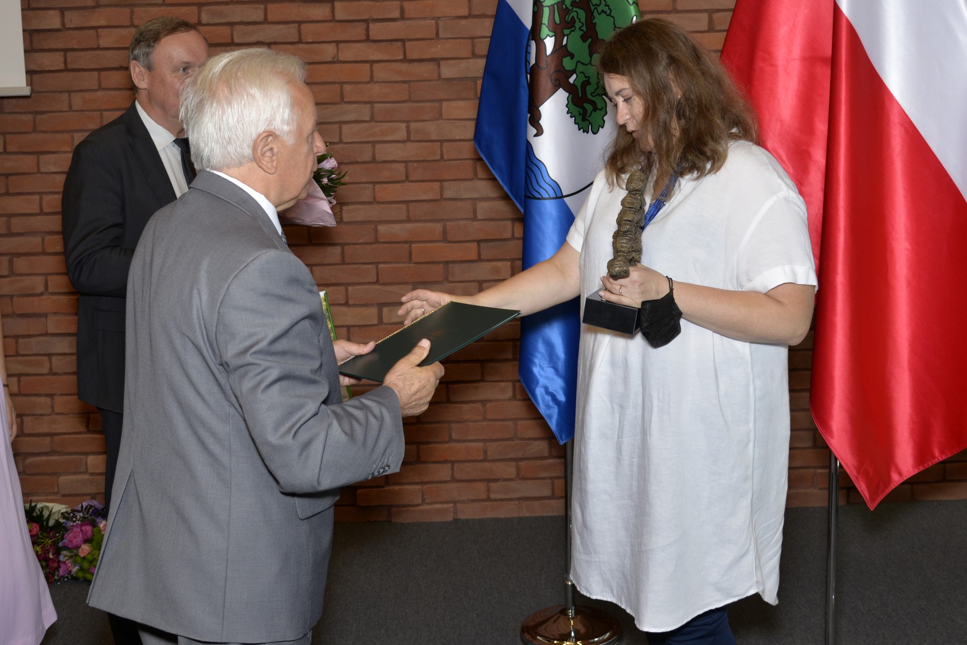 Trzy osoby w sali posiedzeń. Mężczyzna w szarym garniturze przekazuje zieloną teczkę kobiecie trzymającej statuetkę w kształcie dębu. Na drugim planie mężczyzna w czarnym garniturze trzyma w ręku kwiaty.