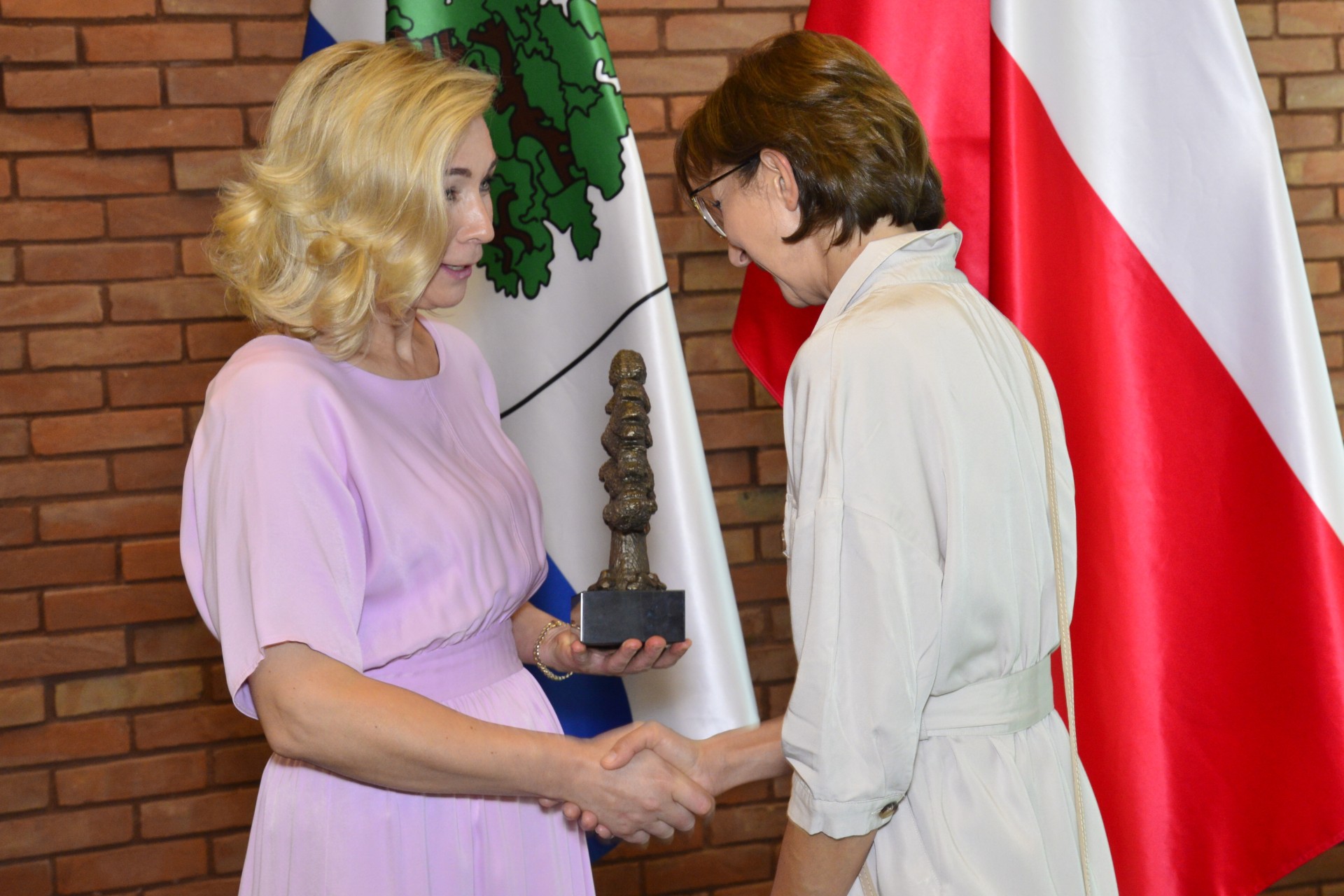 Kobieta w różowej sukience przekazuje drugiej kobiecie statuetkę w kształcie dębu i ściska jej dłoń w geście gratulacji.