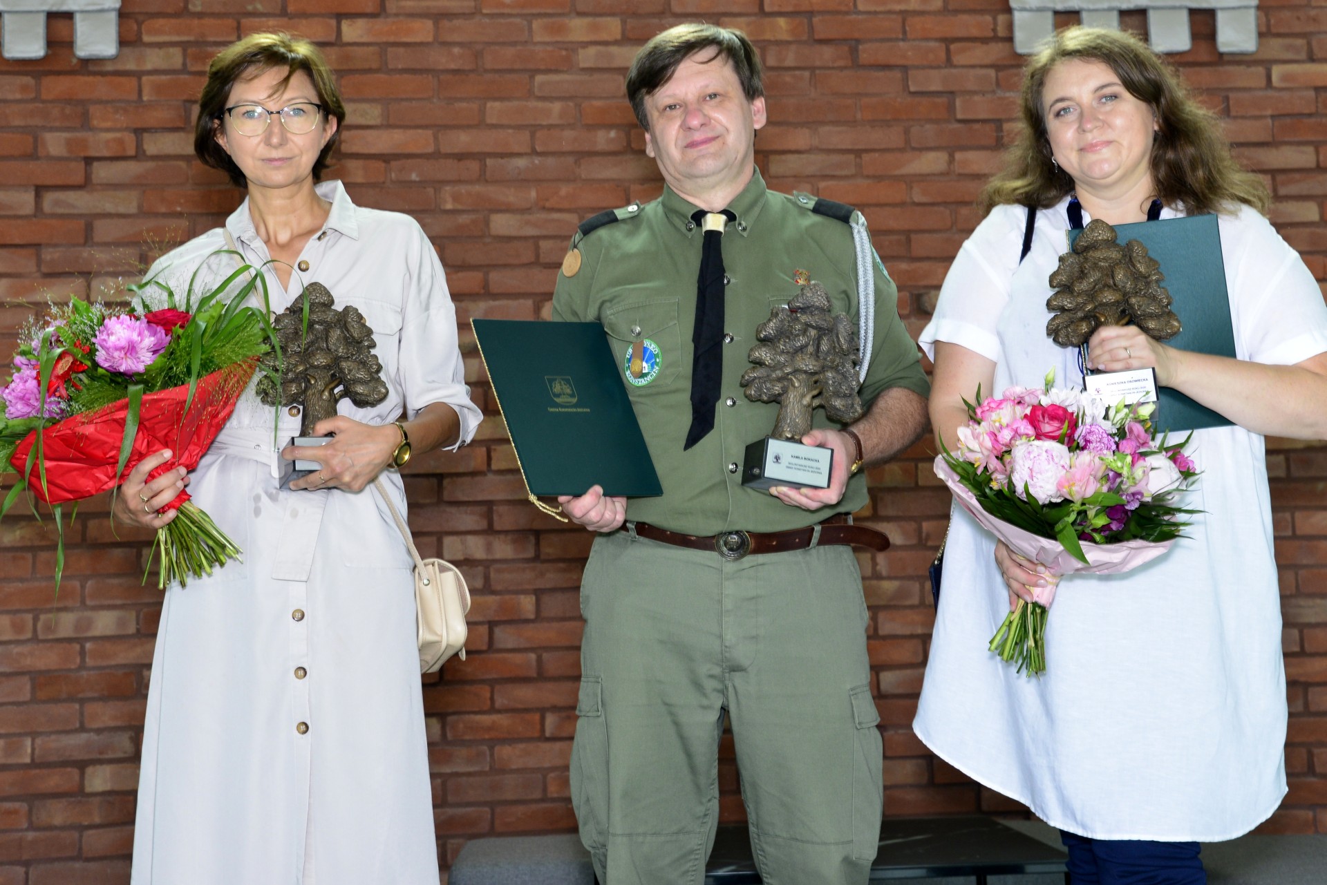 Trzy osoby – dwie kobiety i mężczyzna. Kobiety trzymają w rękach po bukiecie kwiatów i statuetce w kształcie dębu, pomiędzy nimi stoi mężczyzna w mundurze harcerskim trzymający w jednej ręce teczkę z dyplomem, a w drugiej – statuetkę dębu.