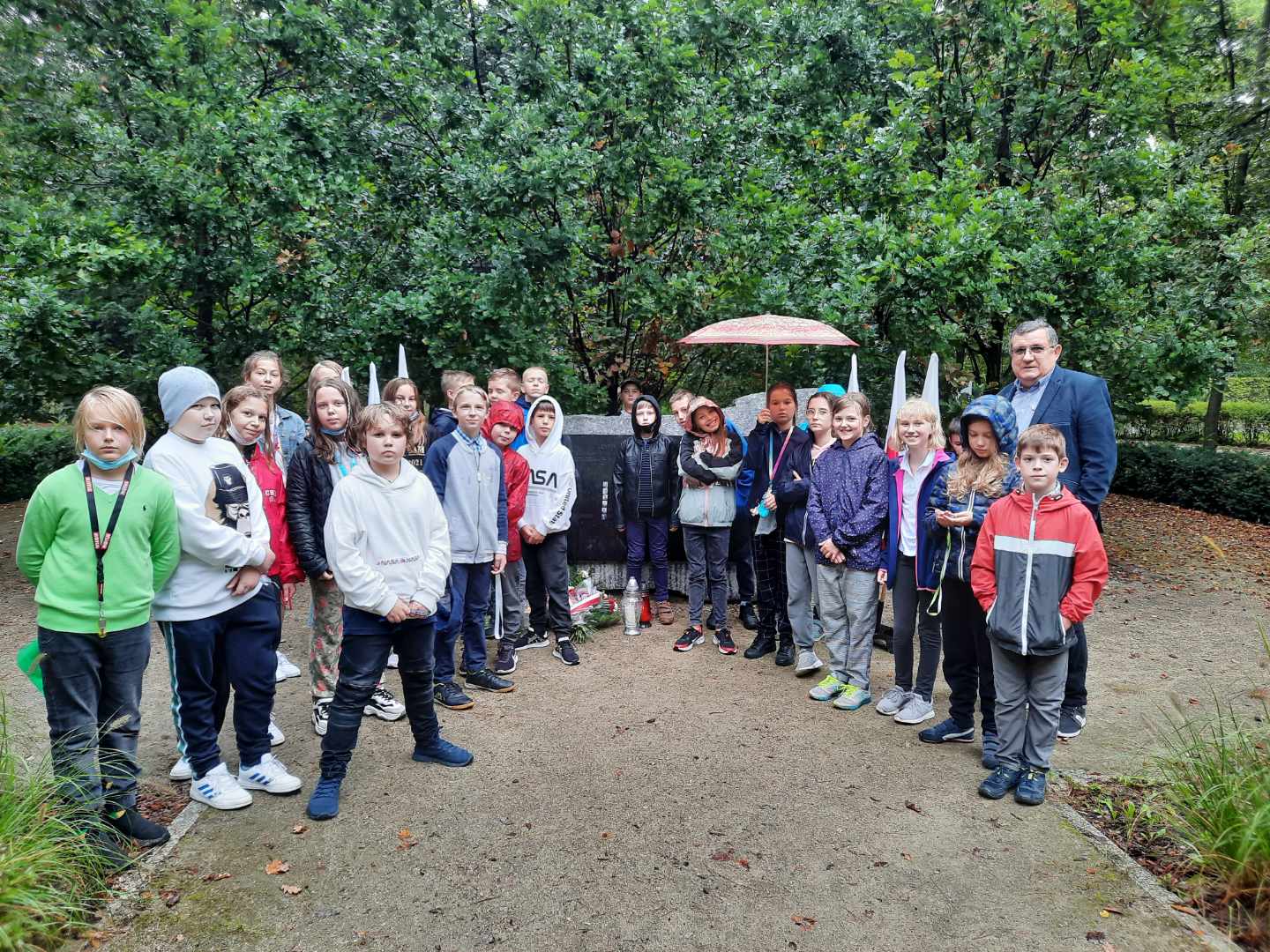 Park. Przy pomniku ofiar katyńskich stoi około trzydzieści osób – grupa dzieci w wieku szkolnym i jeden mężczyzna w średnim wieku. Przy pomniku leży wiązanka z białymi kwiatami i stoją trzy znicze. W tle zielone drzewa.