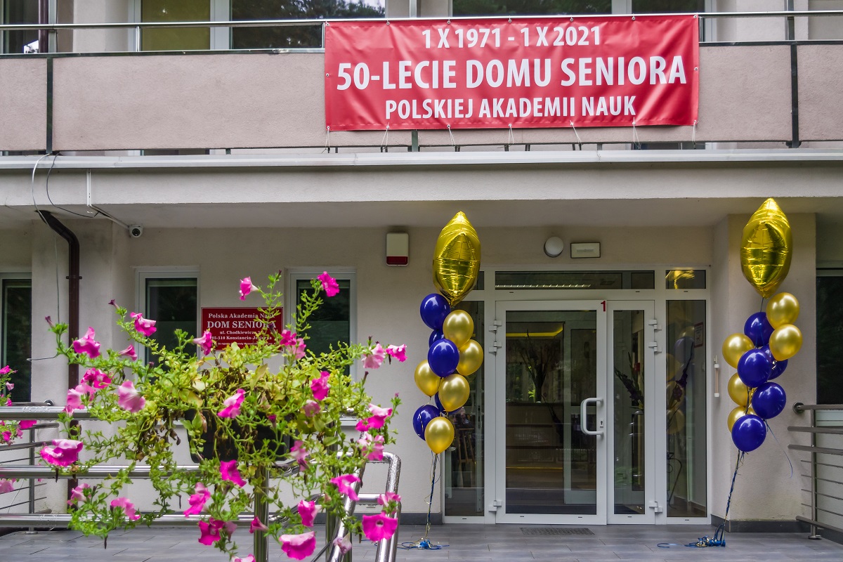 Beżowy budynek. Przed drzwiami po obu stronach są żółto niebieskie balony – ok. 20 sztuk. Obok na budynku przyczepiona jest czerwona tabliczka, na niej są białe napisy Polska Akademia Nauk Dom Seniora. Na górze do balkonu przyczepiony jest czerwony baner, na nim są białe napisy 50-lecie Domu Seniora Polskiej Akademii Nauk.