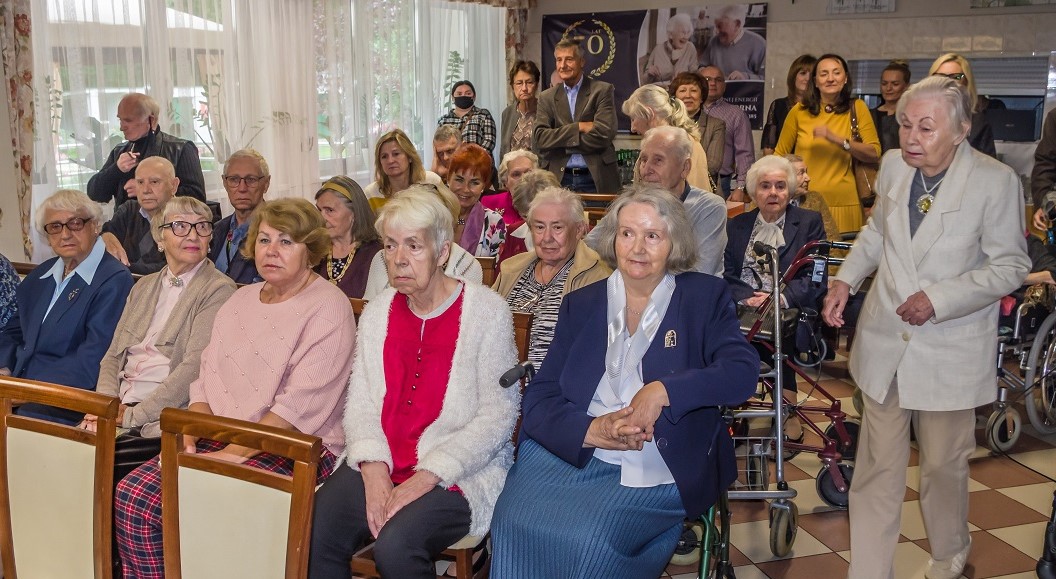 Sala. Na krzesłach siedzi grupa około dwudziestu seniorów – kobiety i mężczyźni. Za nimi stoi około dziesięć osób w średnim wieku. 