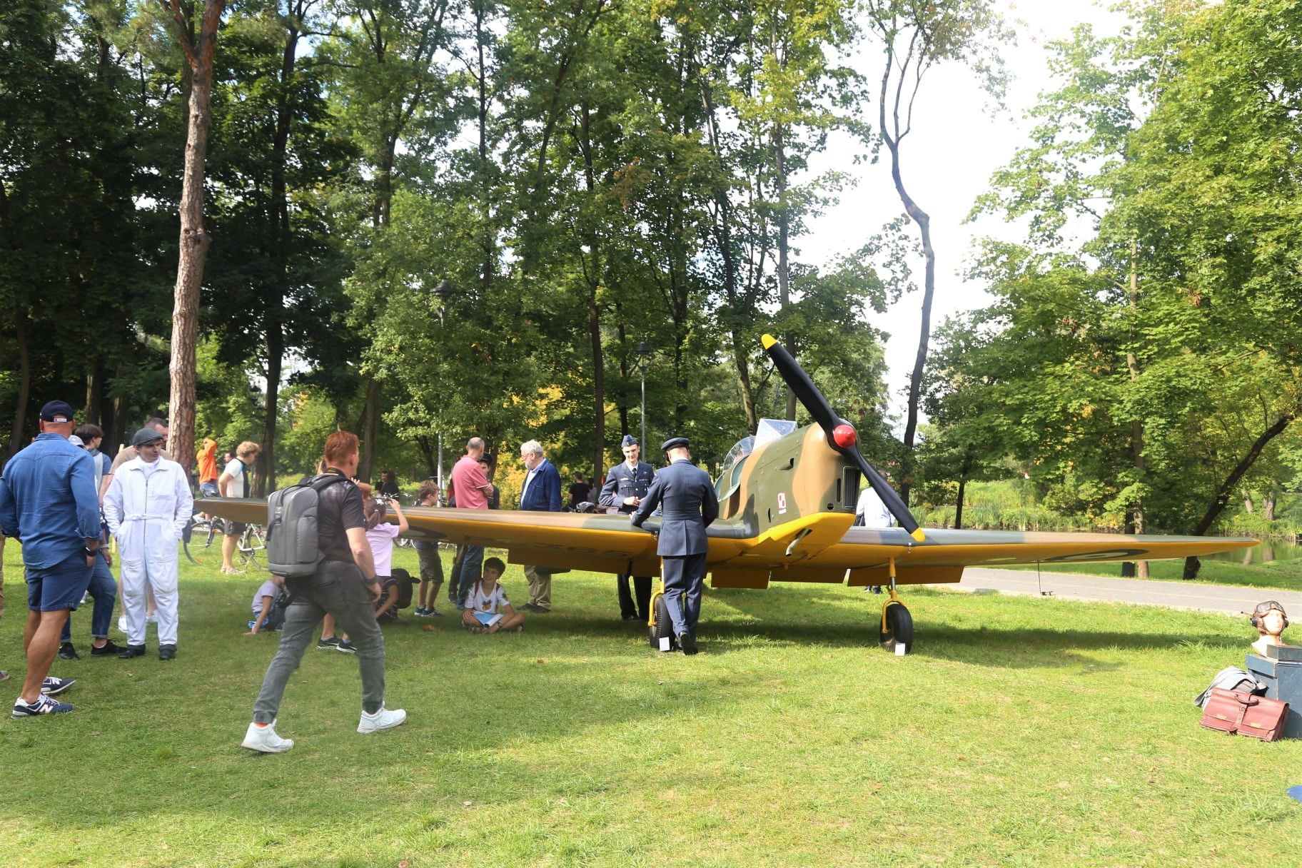 Park Zdrojowy. Na trawie stoi zabytkowy samolot. Dookoła niego stoi około dziesięć osób. Przed nim stoi mężczyzna ubrany w strój pilota, na głowie ma czapkę.