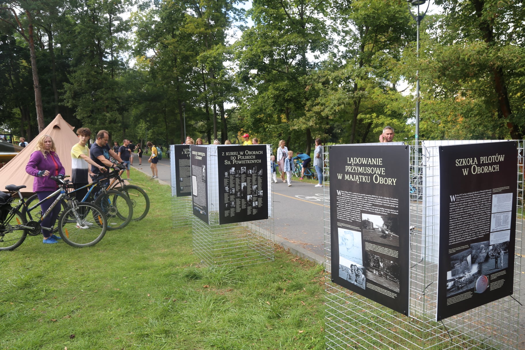 Park Zdrojowy. Na trawie troją trzy tablice informacyjne z historią lotnictwa, na jednej tablicy jest napis szkoła pilotów w oborach, niżej tekst i zdjęcia. W tle są spacerujący alejkami ludzie.