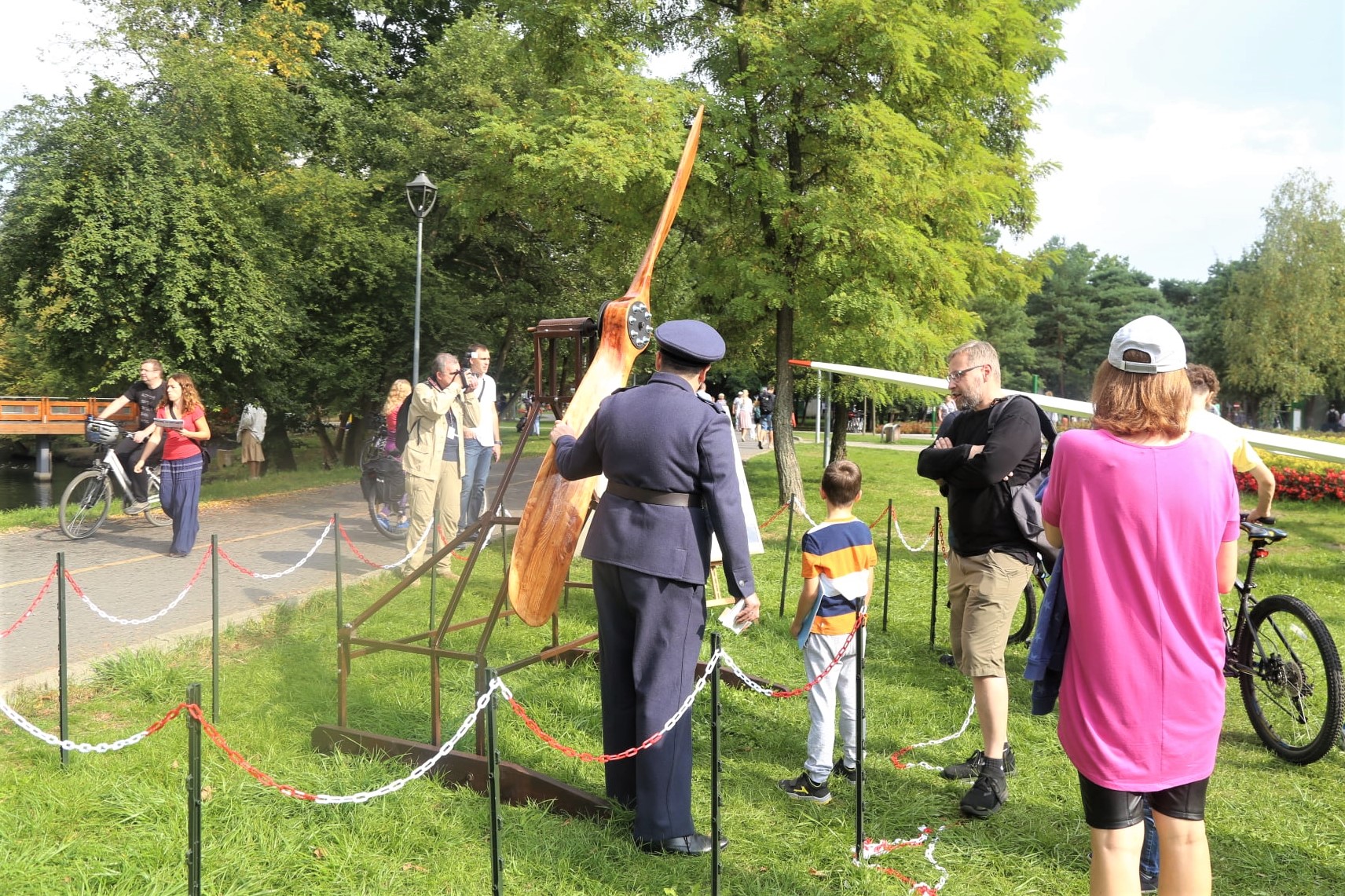 Park Zdrojowy. Na trawie stoi duże drewniane śmigło. Obok stoi mężczyzna ubrany w strój pilota, na głowie ma czapkę. Za nim stoją kobieta i mężczyzna oraz dwójka dzieci.
