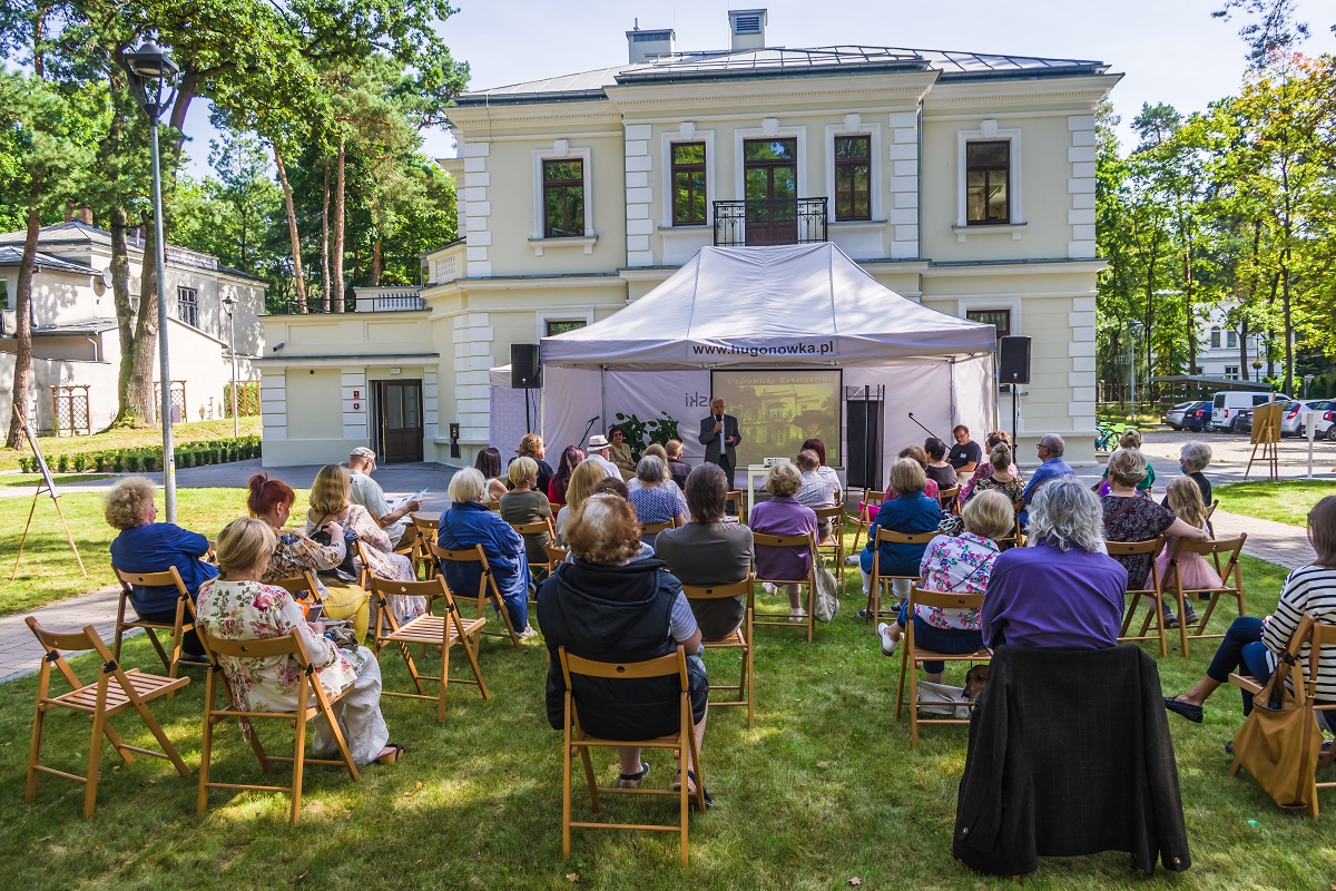 Grupa około trzydziestu osób siedzi na krzesłach. Przed nimi pod namiotem stoi mężczyzna w średnim wieku, ubrany w garnitur, w rękach trzyma mikrofon. W tle jasny budynek.