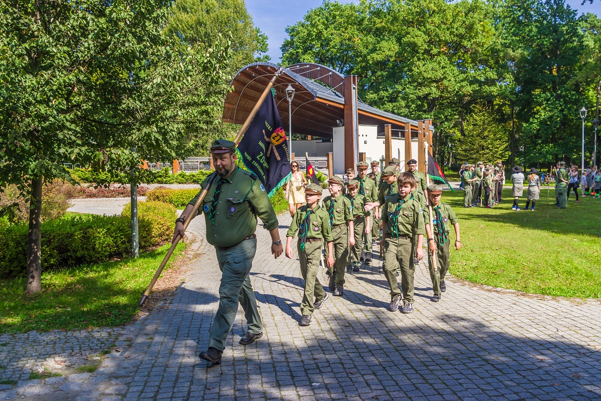 Park Zdrojowy. Mężczyzna ubrany w strój harcerza idzie po chodniku aleją. W ręku trzyma flagę z nazwą grupy harcerskiej. Za nim idzie grupa około dziesięciu osób – dzieci w wieku szkolnym i młodzież. Wszyscy są ubrani w harcerskie stroje. W tle amfiteatr i zielone drzewa.