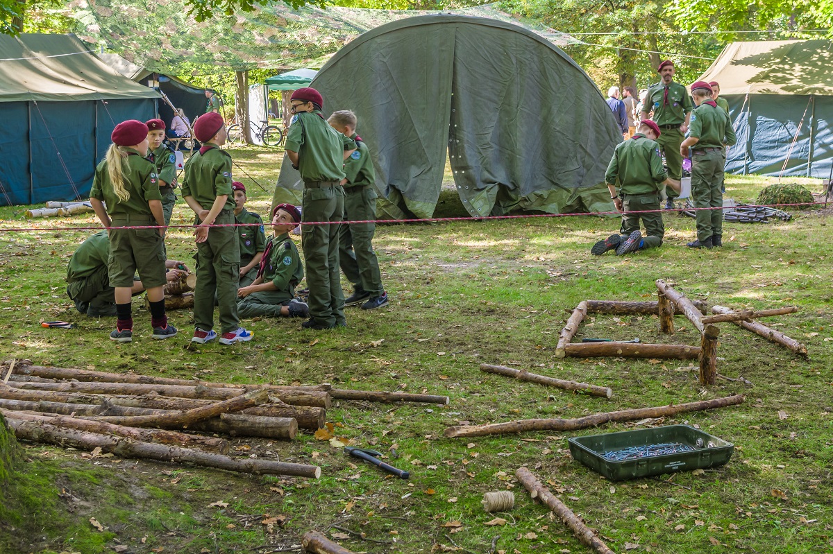  Park Zdrojowy. Po lewej stronie ośmioro dzieci w wieku szkolnym. Stoją obok siebie w grupie. Wszyscy ubrani są w harcerskie stroje, na głowach mają bordowe berety. Przed nimi leżą kawałki drzew. Po prawej stronie stoją cztery osoby – trzech chłopców i jeden mężczyzna. Ubrani są w harcerskie stroje. W tle duży zielony namiot.