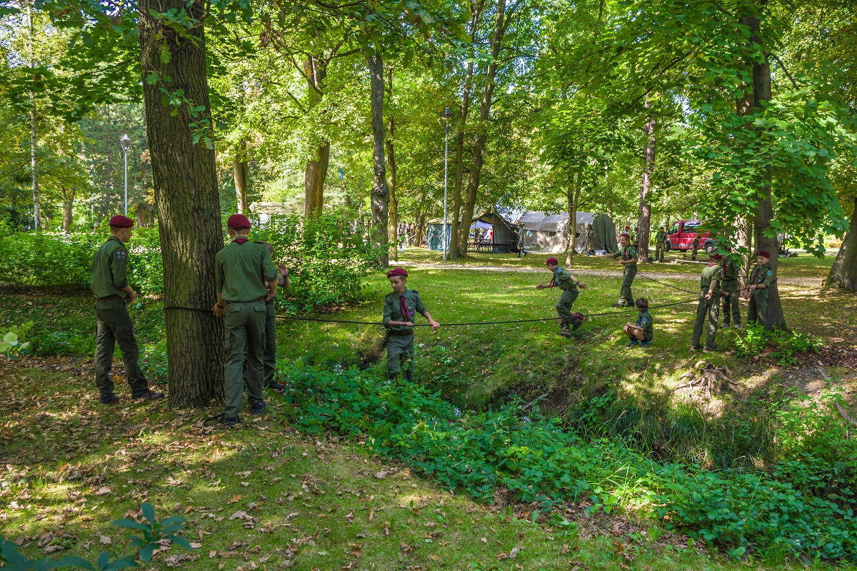 Park Zdrojowy. Grupa dziesięciu osób – dzieci w wieku szkolnym. Stoją obok siebie, rękach trzymają czarną linę, która jest przywiązana do dwóch drzew oddalonych od siebie. Wszyscy ubrani są w harcerskie stroje, na głowach mają bordowe berety.