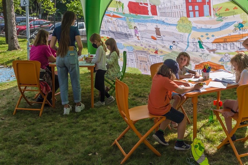 Na trawie pod namiotem stoją dwa stoliki. Przy nich na krzesłach siedzą dzieci w wieku szkolnym. Na stole leżą kartki do kolorowania oraz kredki.