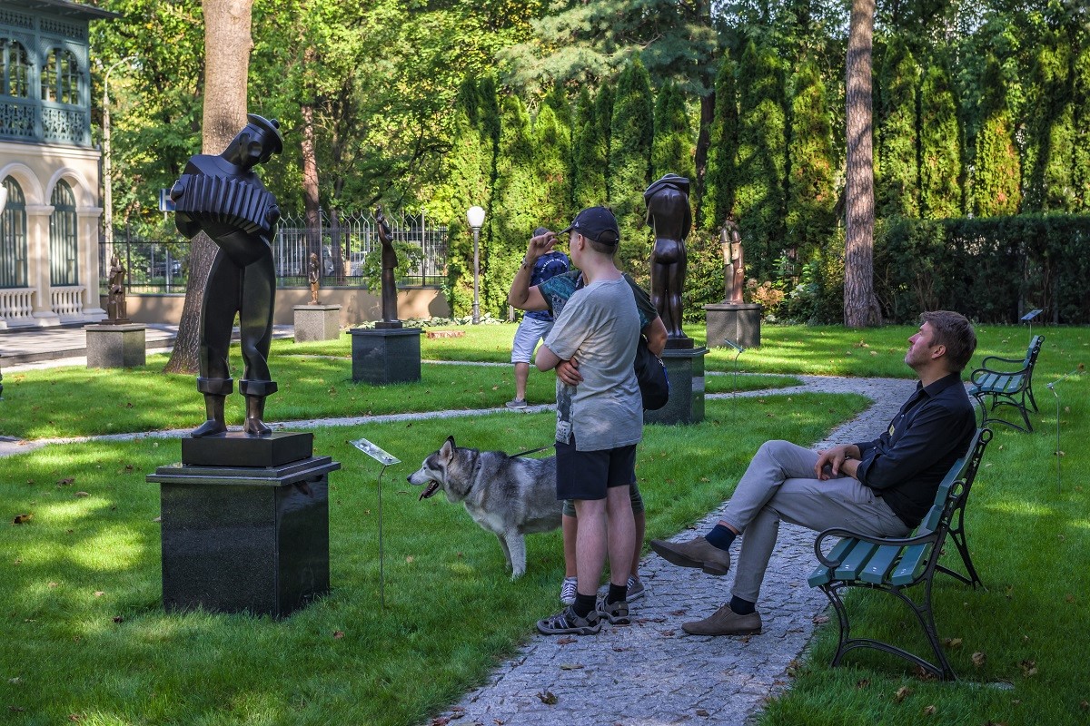 Park rzeźb. Przed rzeźbą na chodniku stoją dwie osoby, obok stoi pies, za nimi na ławce siedzi mężczyzna. W tle alejki z rzeźbami i zielone drzewa.
