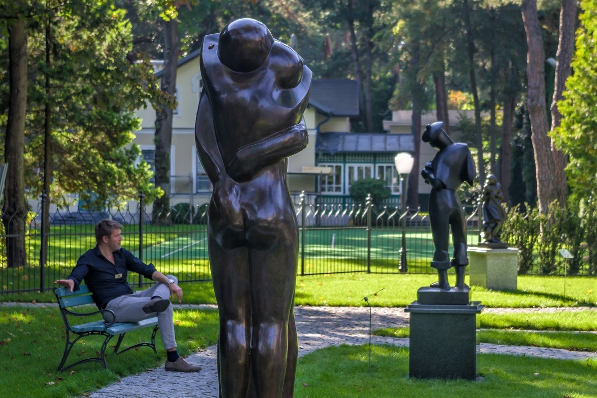 Park rzeźb. Na pierwszym planie brązowa rzeźba przedstawiająca dwie osoby przytulające się do siebie. W tle na ławce przy alejce siedzi mężczyzna, naprzeciwko niego stoi czarna rzeźba. W tle budynek i zielone drzewa.