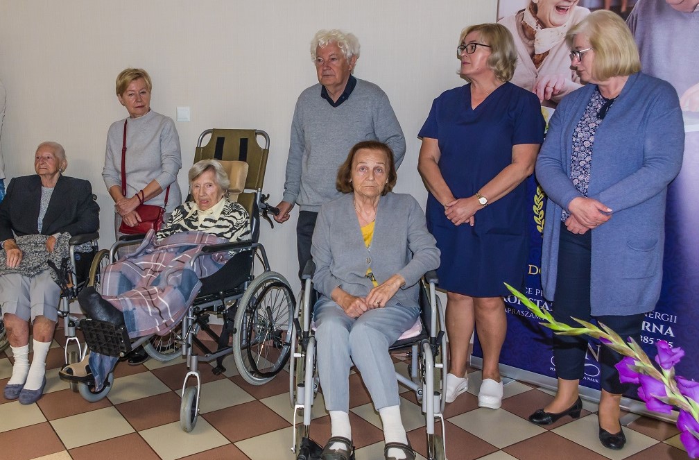 Sala. Trzy kobiety – seniorki siedzą na wózkach inwalidzkich. Obok stoją cztery osoby trzy kobiety w średnim wieku oraz mężczyzna – senior.