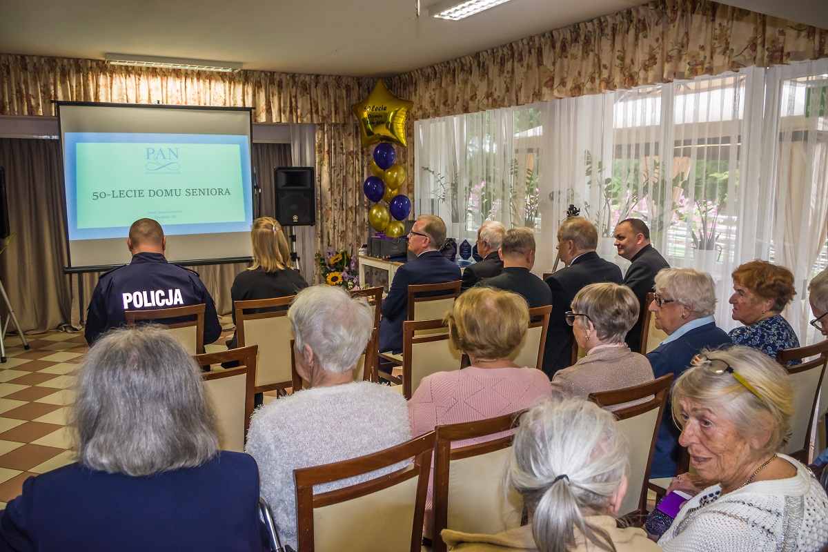 Sala. Około dwadzieścia osób siedzi na krzesłach. Z tyłu siedzą seniorzy. Z przodu siedzi kobieta oraz sześciu mężczyzn w średnim wieku. Jeden z nich ma kurtkę z napisem Policja.  Naprzeciwko nich jest projektor na którym jest wyświetlana prezentacja o Domu Seniora.