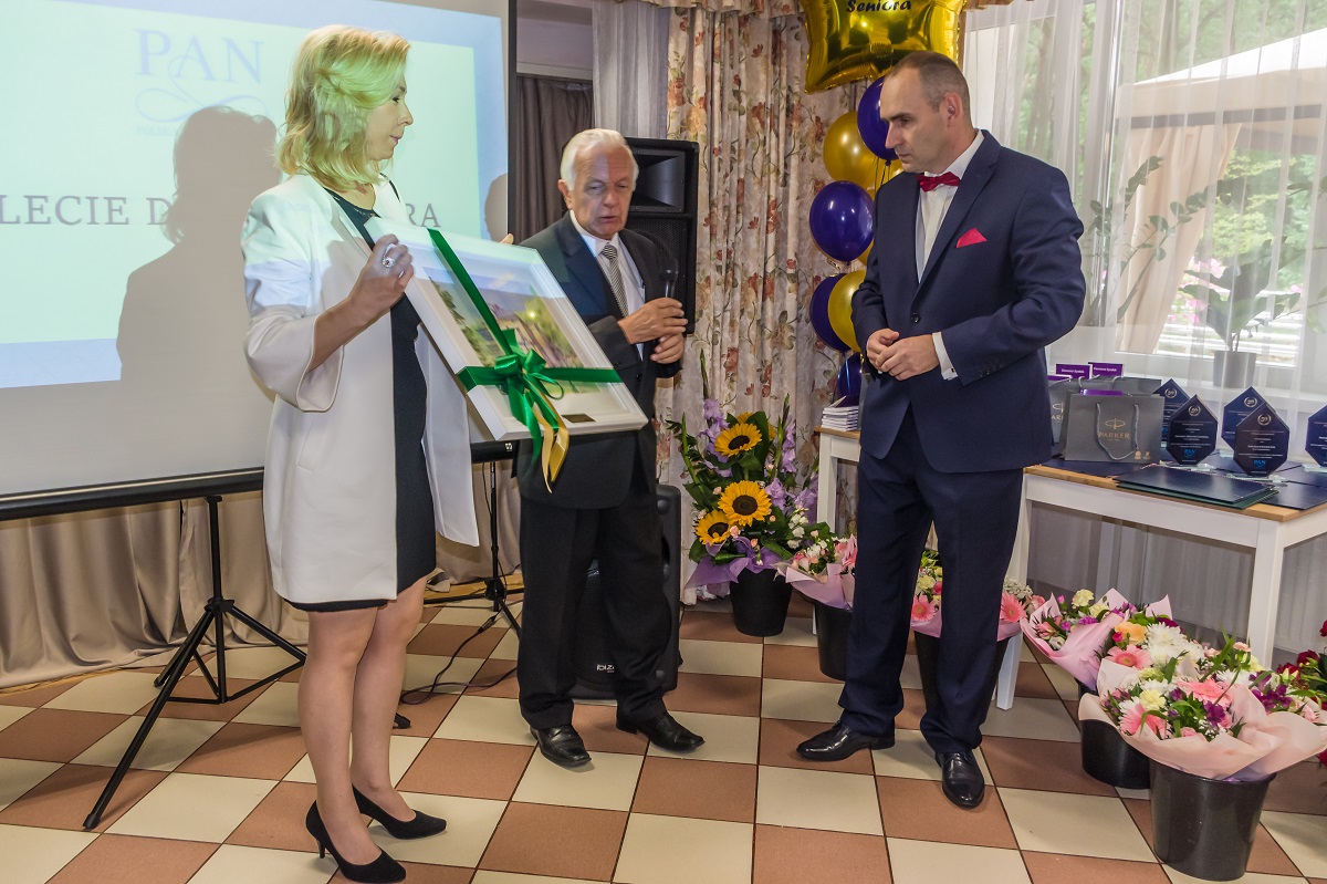 Sala. Trzy osoby stoją obok siebie – dwóch mężczyzn i kobieta. Mężczyźni ubrani są w garnitury. Kobieta ubrana jest w czarną sukienkę. Kobieta w rękach trzyma ramę, w środku jest obraz namalowany farbami.