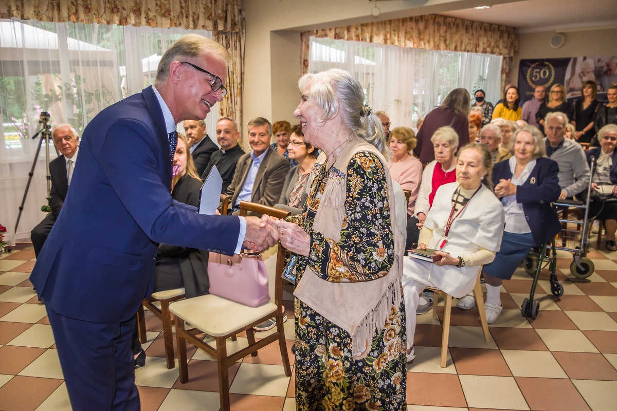 Sala. Mężczyzna w średnim wieku ubrany w garnitur oraz seniorka stoją naprzeciwko siebie, ściskają sobie prawe dłonie. Za nimi na krzesłach siedzi około dwadzieścia osób.