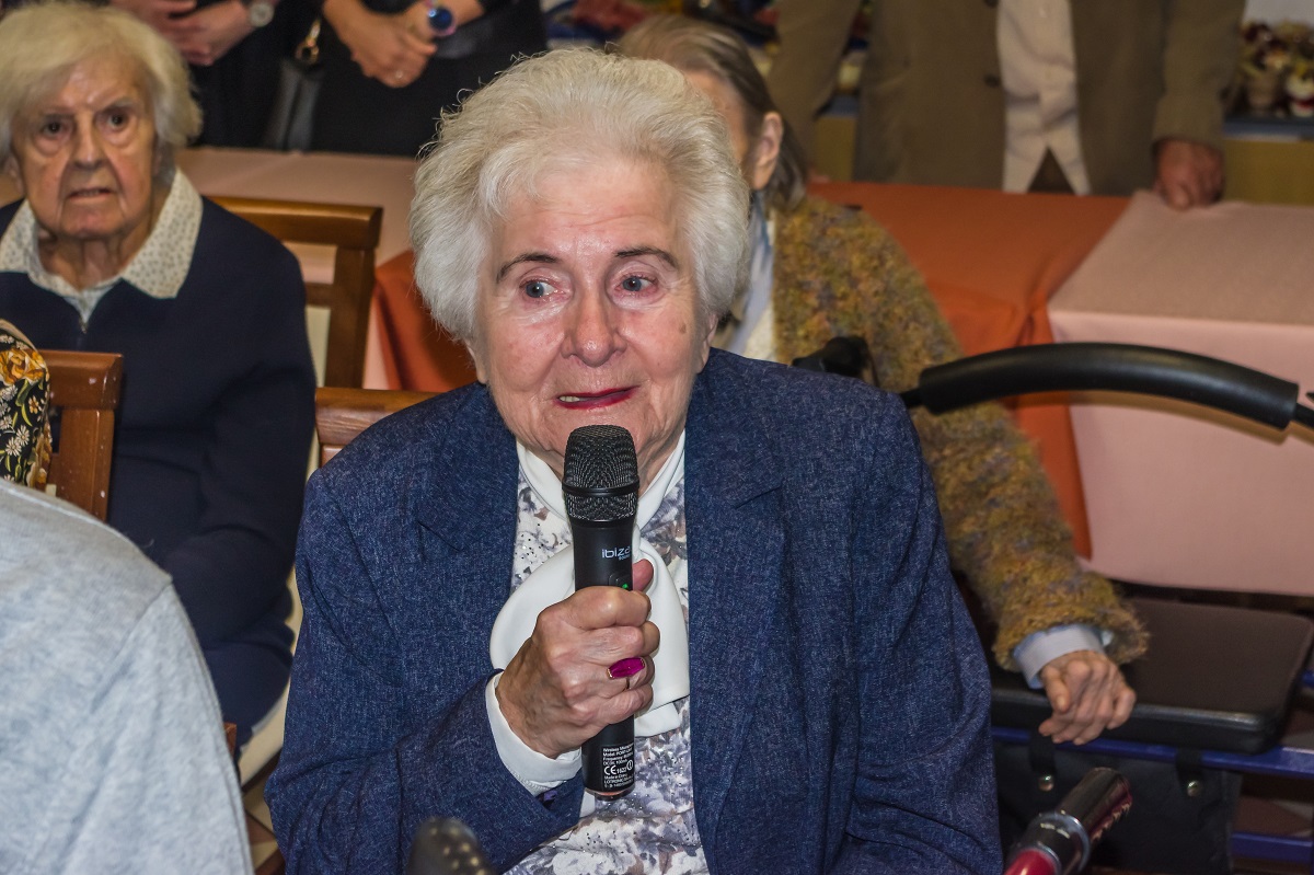 Sala. Seniorka siedzi na krześle, w prawej ręce trzyma mikrofon. Za nią siedzą na krzesłach dwie seniorki.