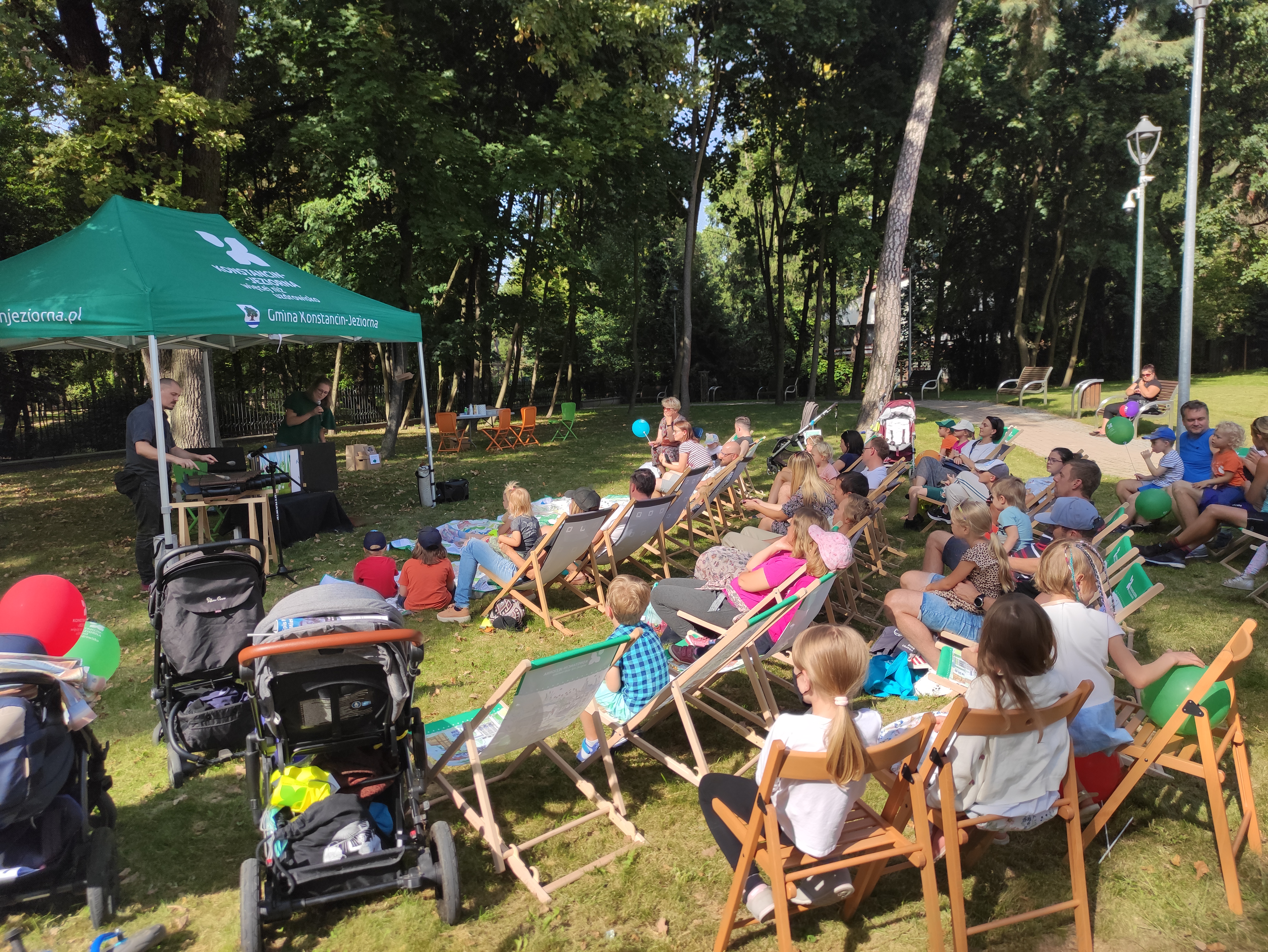 Park Zdrojowy. Po lewej stronie pod zielonym namiotem stoją dwie osoby. Naprzeciwko nich siedzi na krzesłach i na leżakach grupa około trzydziestu osób – dzieci i osoby dorosłe.