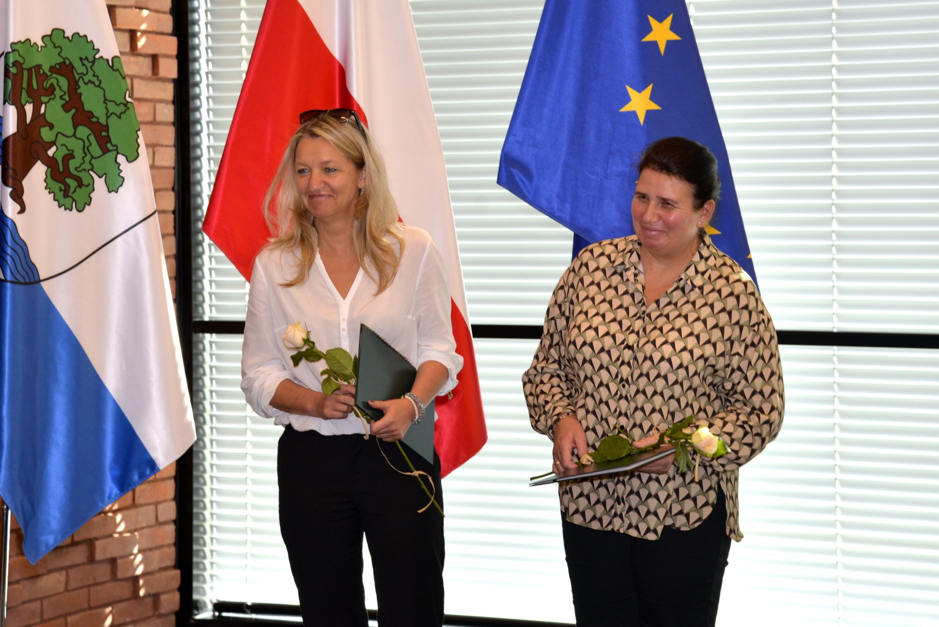 Sala konferencyjna. Dwie kobiety w średnim wieku stoją obok siebie, w rękach trzymają dyplomy i białe róże. W tle trzy flagi: flaga Konstancina-Jeziorny, flaga Polski i flaga Unii Europejskiej oraz okno z żaluzjami.