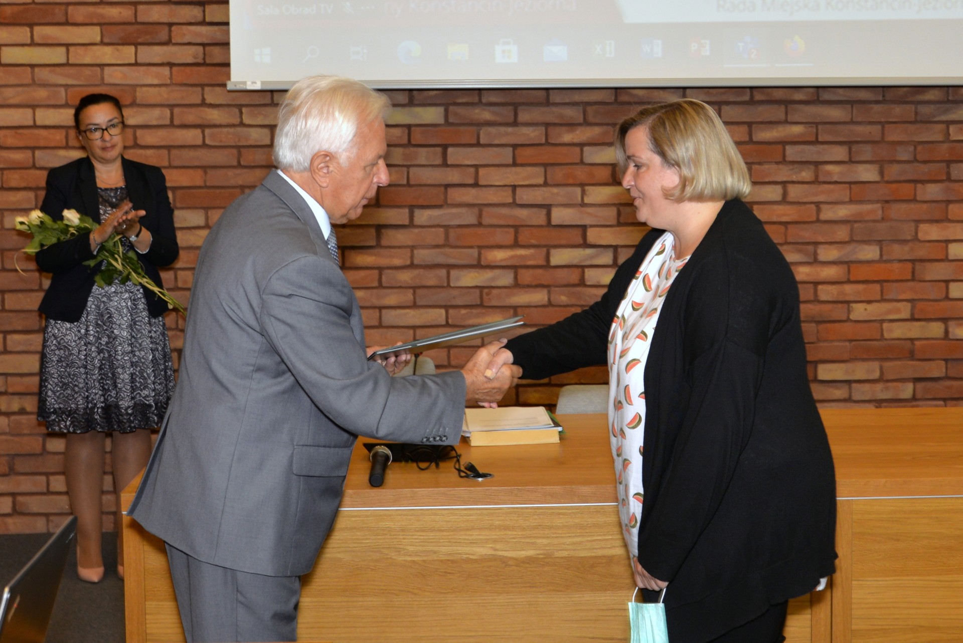 Sala konferencyjna. Mężczyzna w średnim wieku ubrany w garnitur, w rękach trzyma teczkę. Naprzeciwko niego stoi kobieta, która ubrana jest w białą bluzkę i czarny sweter. Ściskają sobie prawe dłonie. W tle ściana z czerwonej cegły.