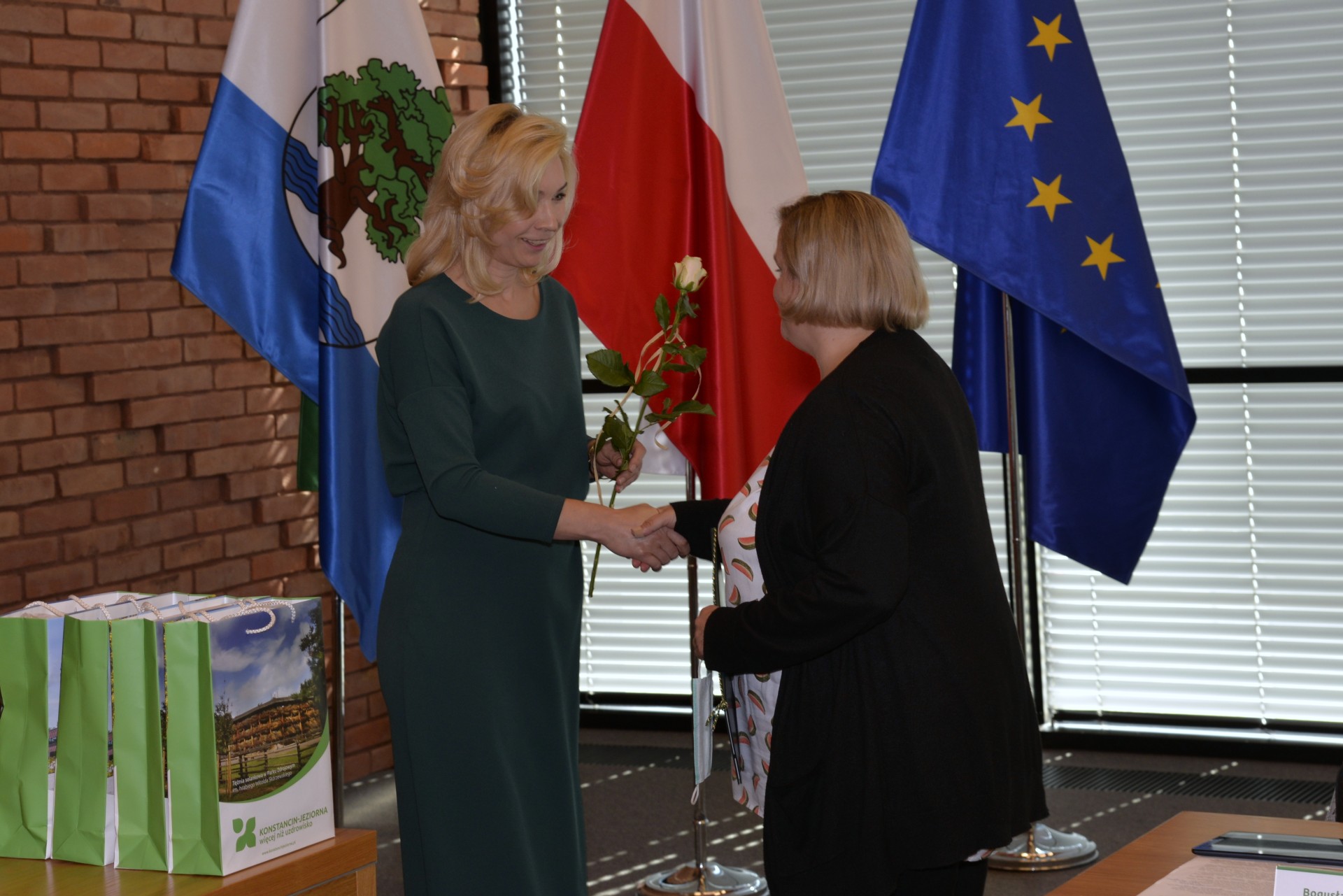 Sala konferencyjna. Dwie kobiety stoją naprzeciwko siebie. Ściskają sobie prawe dłonie. Jedna z kobiet w ręku trzyma białą różę. Obok na stoliku stoją cztery torby prezentowe. W tle trzy flagi: flaga Konstancina-Jeziorny, flaga Polski i flaga Unii Europejskiej.