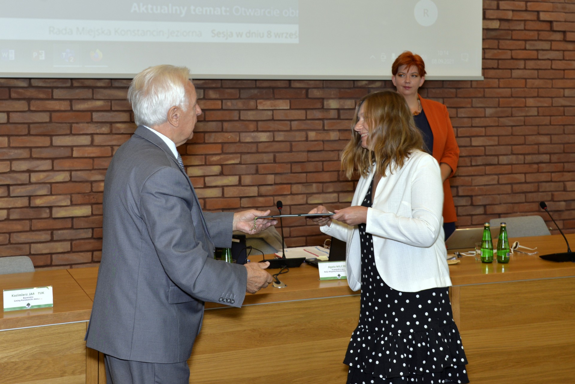 Sala konferencyjna. Mężczyzna w średnim wieku ubrany w garnitur.  Naprzeciwko niego stoi kobieta, która ubrana jest w czarną sukienkę i białą marynarkę. Mężczyzna wręcza kobiecie dyplom. W tle ściana z czerwonej cegły.