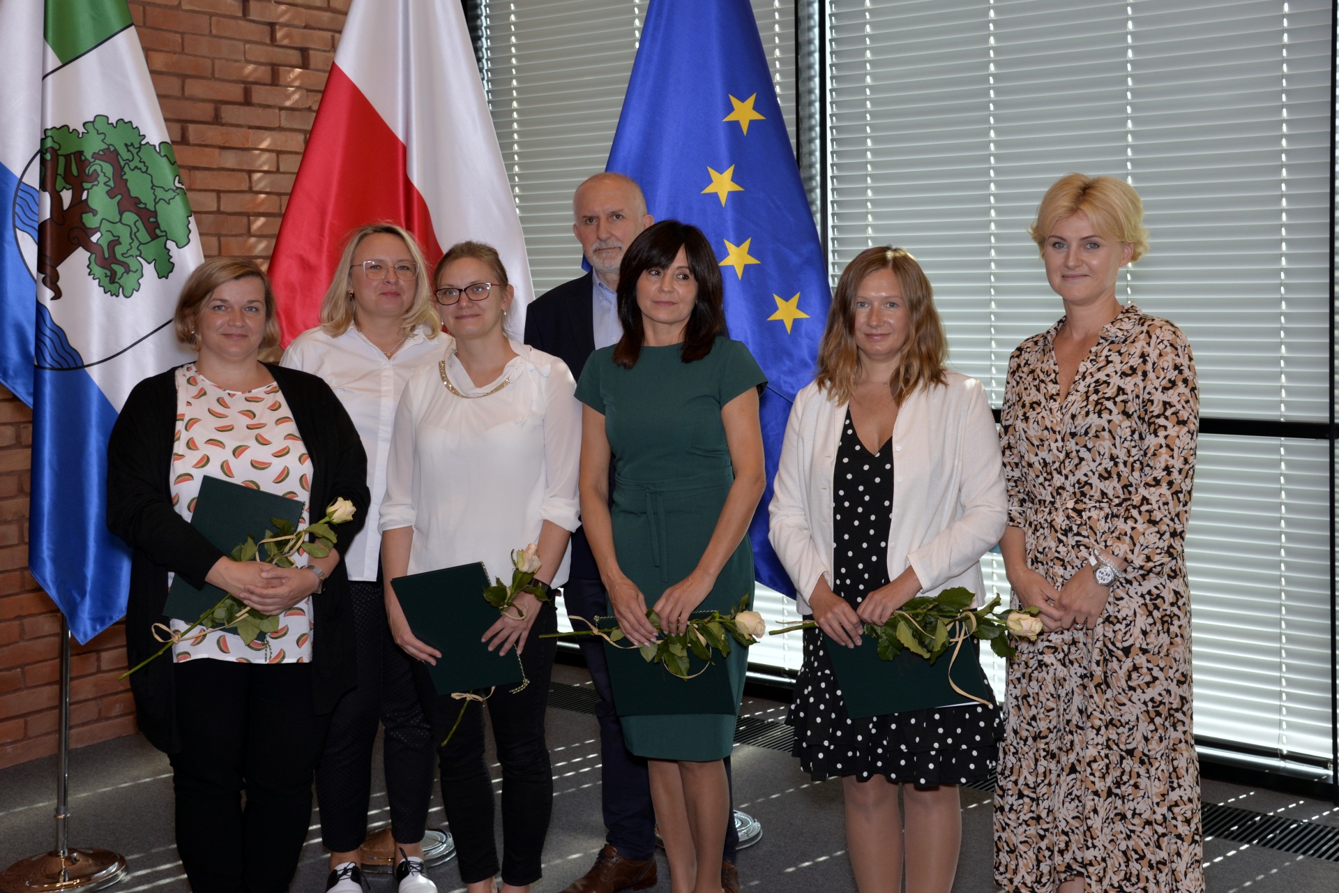 Sala konferencyjna. Siedem osób stoi obok siebie w rzędzie – sześć kobiet i jeden mężczyzna. Cztery kobiety w rękach trzymają dyplomy i białe róże. W tle trzy flagi: flaga Konstancina-Jeziorny, flaga Polski i flaga Unii Europejskiej.