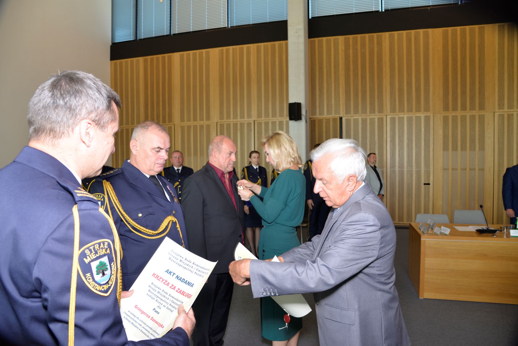 Sala konferencyjna. Mężczyzna w średnim wieku, ubrany w garnitur, w rękach trzyma dyplom. Naprzeciwko niego stoi dwóch mężczyzn, którzy ubrany są w granatowe mundury Straży Miejskiej. Za nim stoją kobieta i mężczyzna. Kobieta wręcza coś mężczyźnie. 