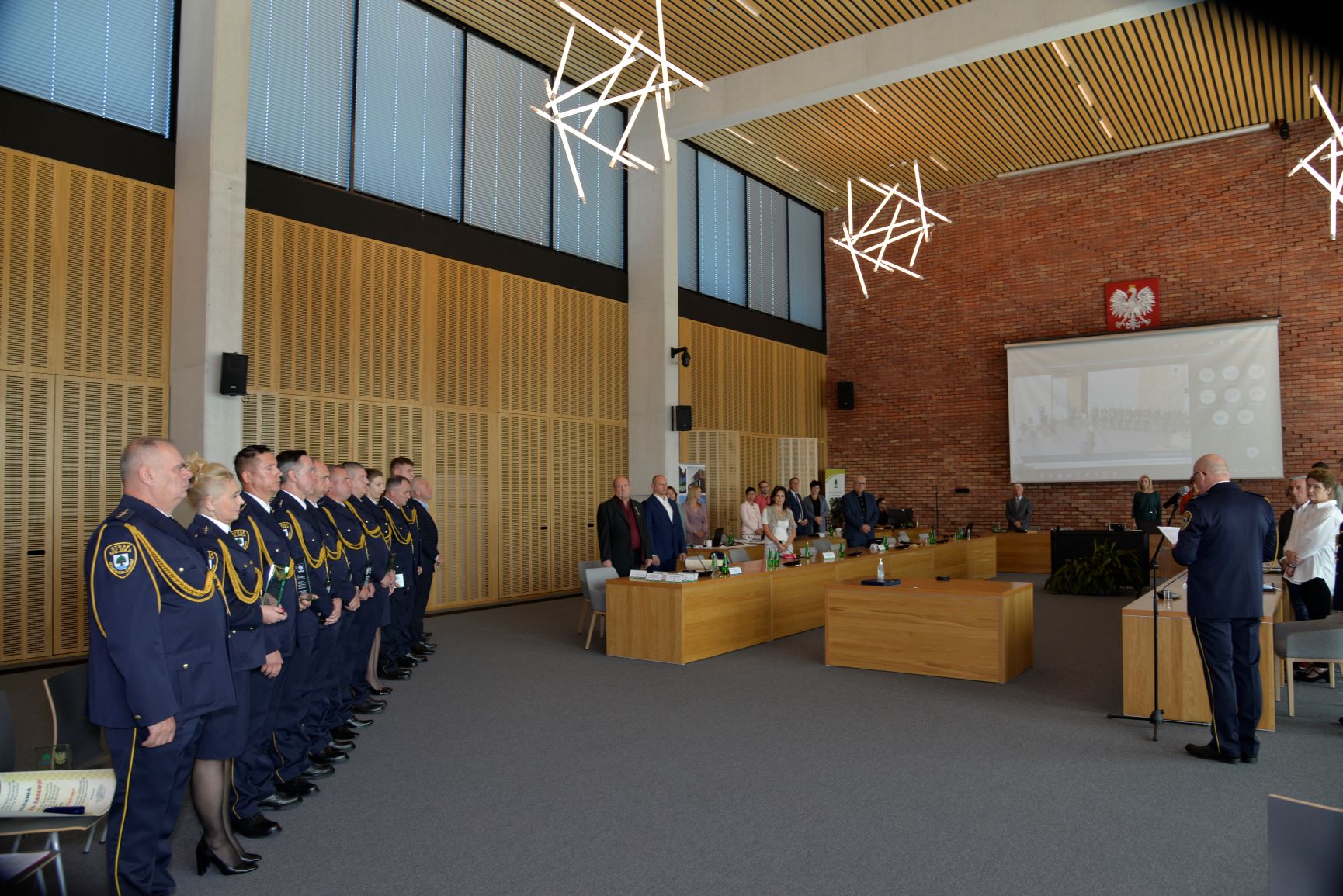 Sala konferencyjna. Po lewej stronie stoi obok siebie w rzędzie jedenaście osób – dwie kobiety i dziewięciu mężczyzn. Wszyscy ubrani są w granatowe mundury Straży Miejskiej. Po lewej stronie znajdują się trzy drewniane stoły ustawione obok siebie. Przy stołach stoi około dwadzieścia osób.