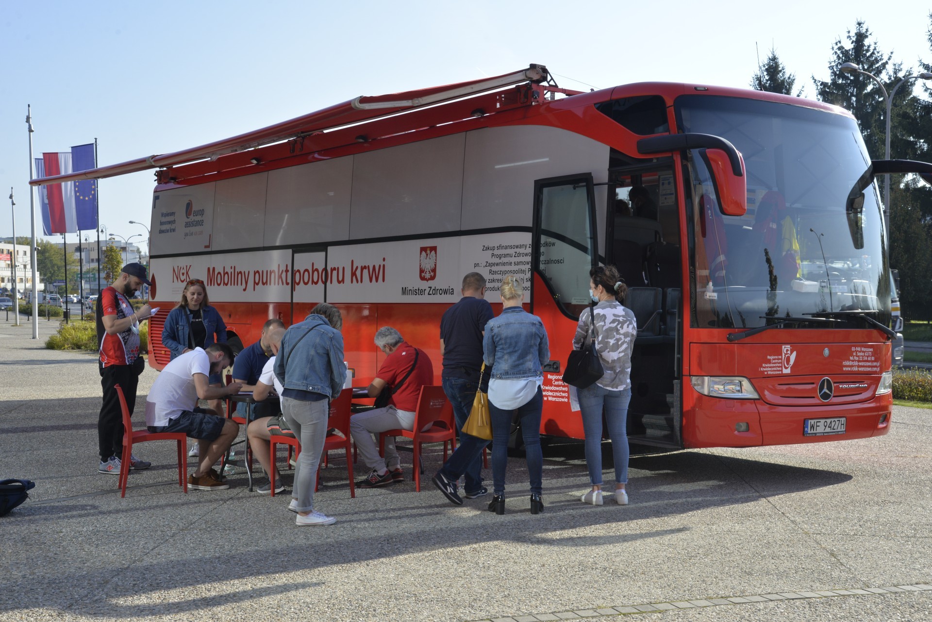 Miasto. Na chodniku stoi czerwony autokar, z boku ma duży napis Mobilny punkt poboru krwi, obok jest godło Polski i napis Minister Zdrowia.  Przed autokarem stoi sześć osób oraz na krzesłach przy stoliku siedzą cztery osoby.