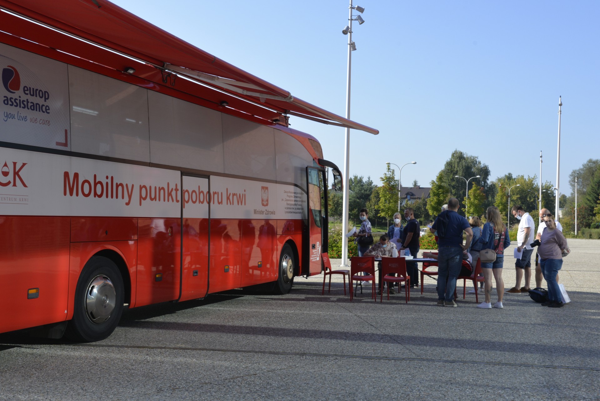 Miasto. Na chodniku stoi czerwony autokar, z boku ma duży napis Mobilny punkt poboru krwi. Przed autokarem w kolejce stoi dziesięć osób. Jedna osoba siedzi na krześle przy stoliku. W tle zielone drzewa i błękitne niebo.