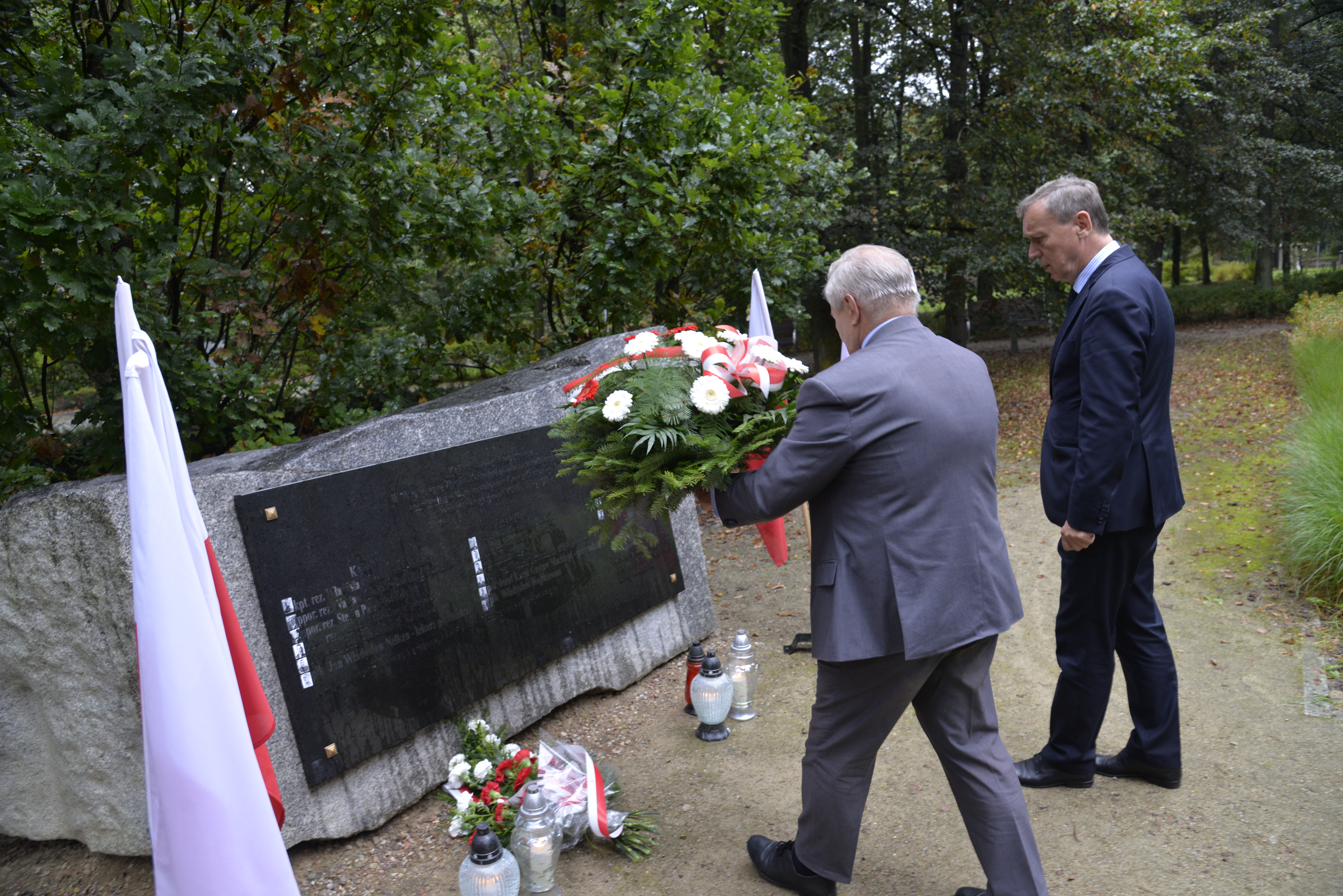 Park. Przed pomnikiem ofiar katyńskich stoi dwóch mężczyzn w średnim wieku. Jeden z nich trzyma wiązankę z białymi kwiatami. Przy pomniku stoją znicze i leżą biało-czerwone kwiaty. Po obu stronach pomnika stoją na stojaku biało-czerwone flagi.