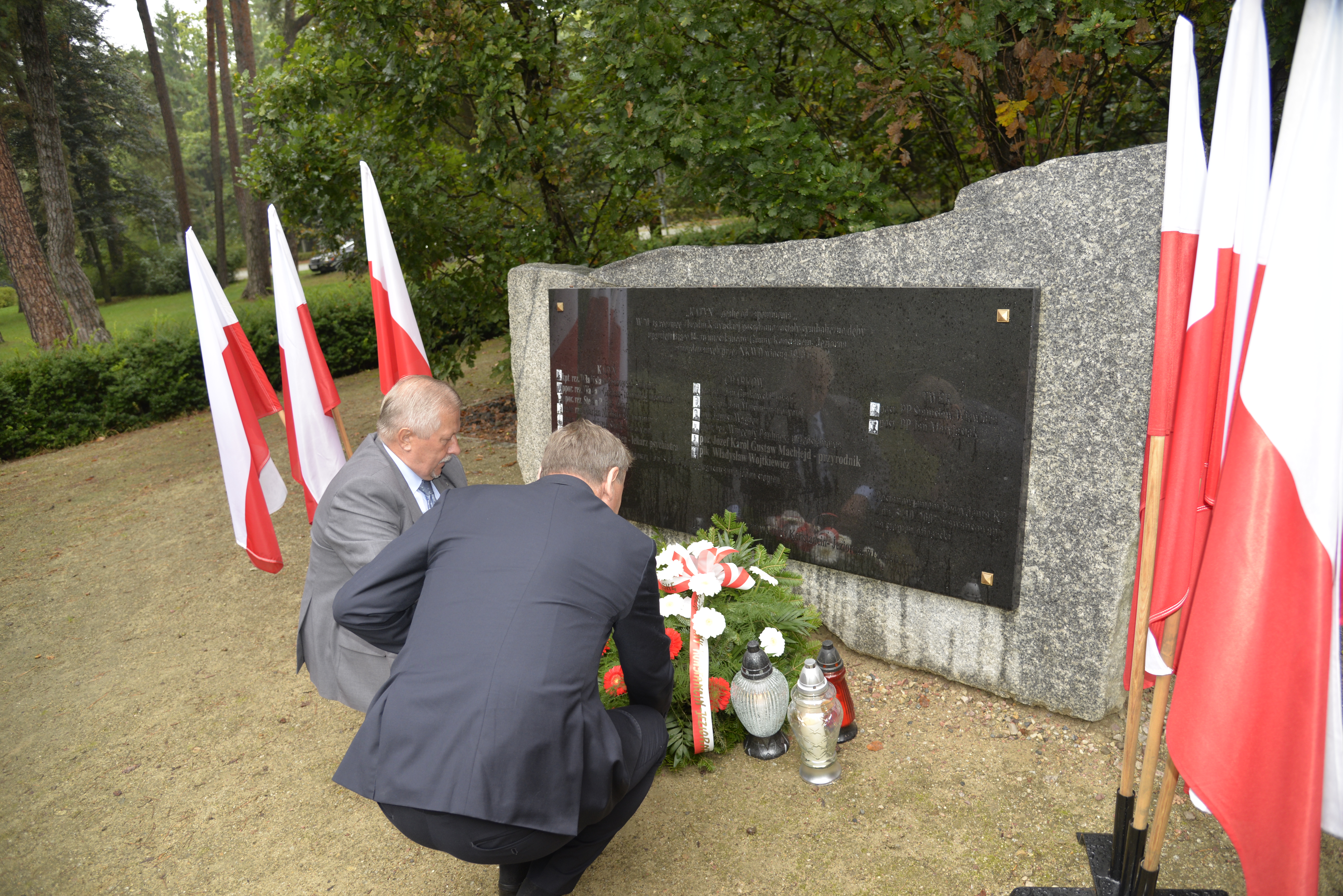 Park. Przed pomnikiem ofiar katyńskich dwóch mężczyzn w średnim wieku kuca. Przy pomniku leży wiązanka z białymi kwiatami i stoją trzy znicze. Po obu stronach pomnika stoją na stojaku trzy biało-czerwone flagi.