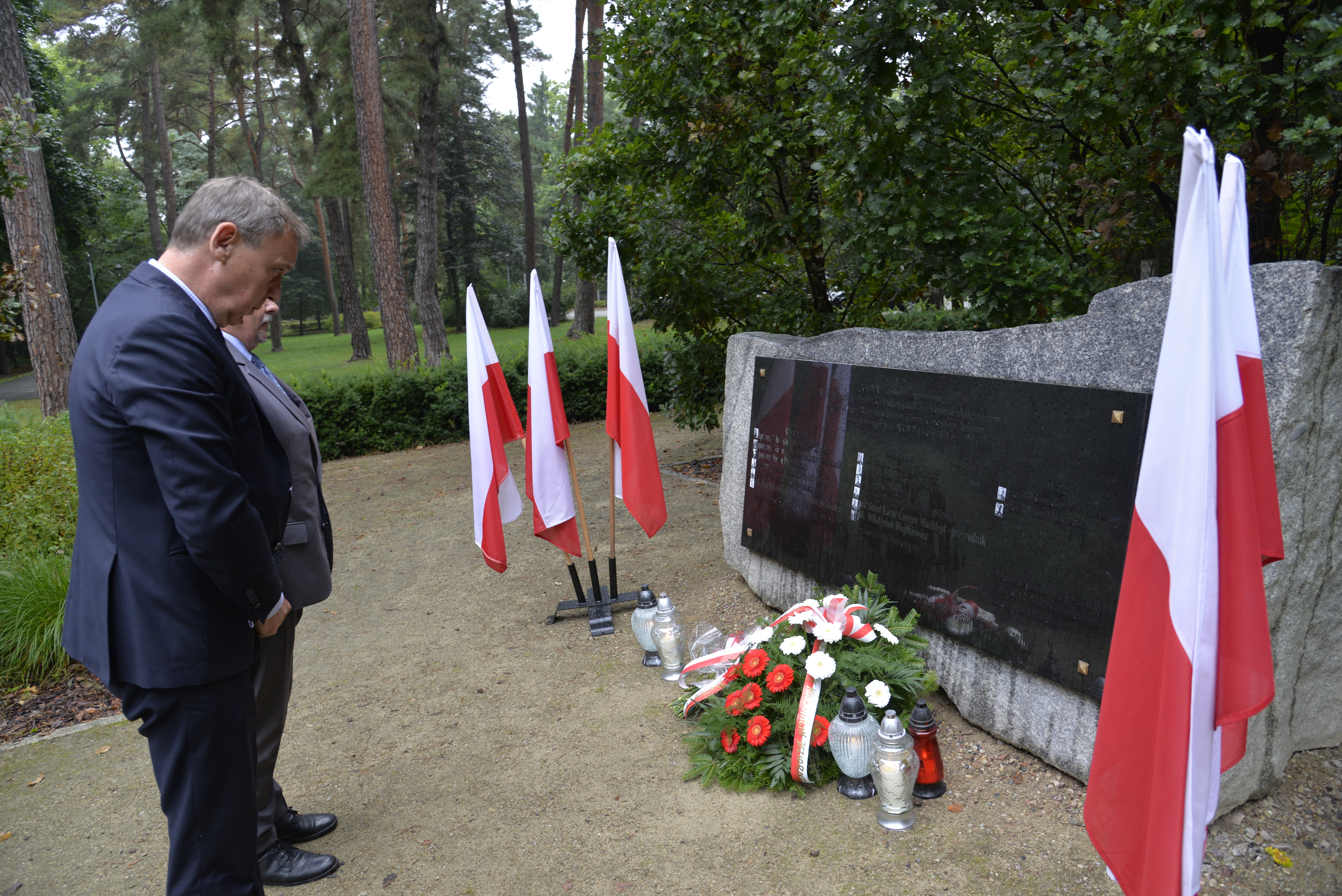 Park. Przed pomnikiem ofiar katyńskich stoi dwóch mężczyzn w średnim wieku. Przy pomniku stoją znicze i leży wiązanka z białymi kwiatami. Po obu stronach pomnika stoją trzy biało-czerwone flagi.