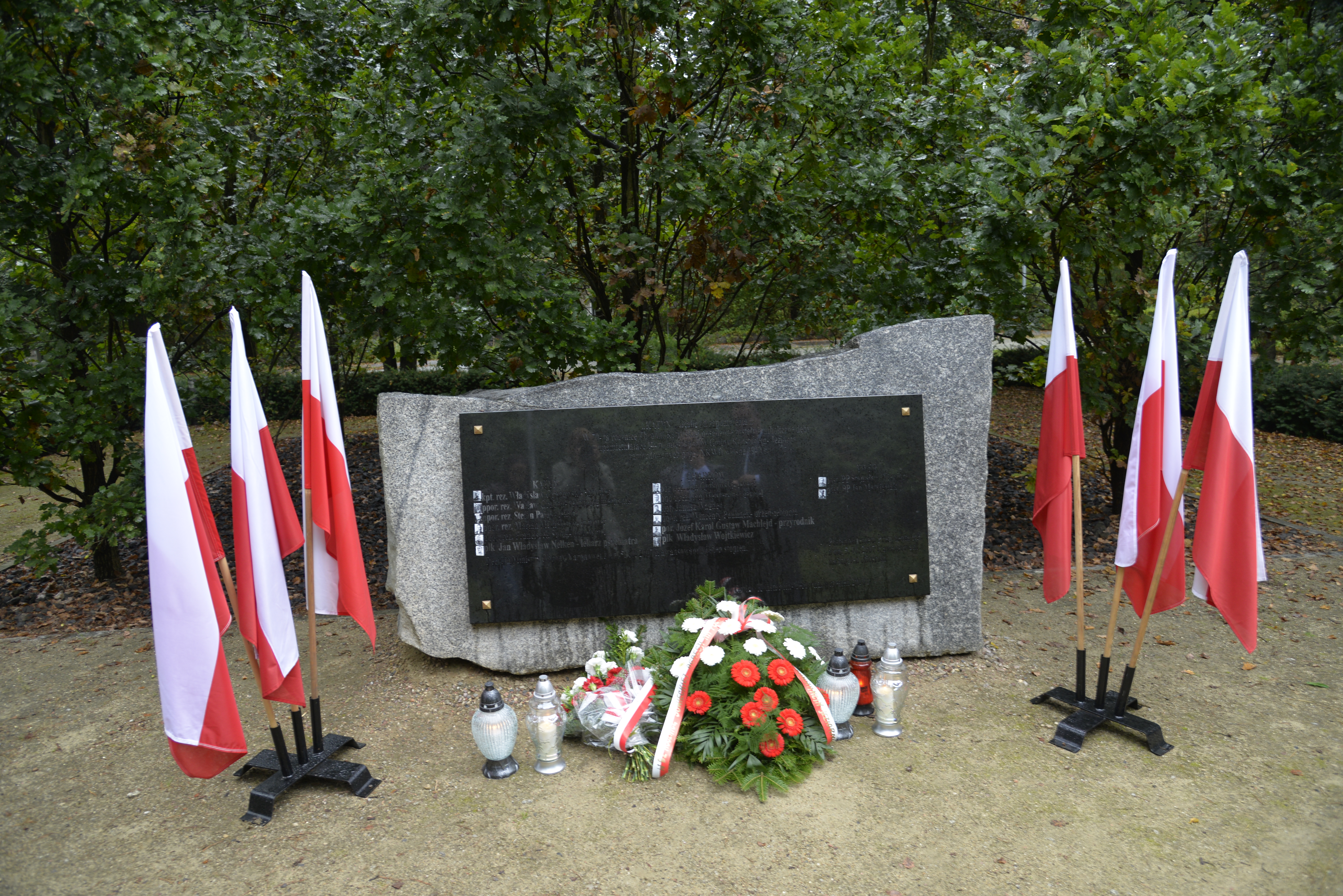 Park. Pomnik ofiar katyńskich. Przy nim stoją znicze oraz leżą: biało-czerwone kwiaty i wiązanka z białymi kwiatami. Po obu stronach pomnika stoją na stojaku trzy biało-czerwone flagi. W tle zielone drzewa.
