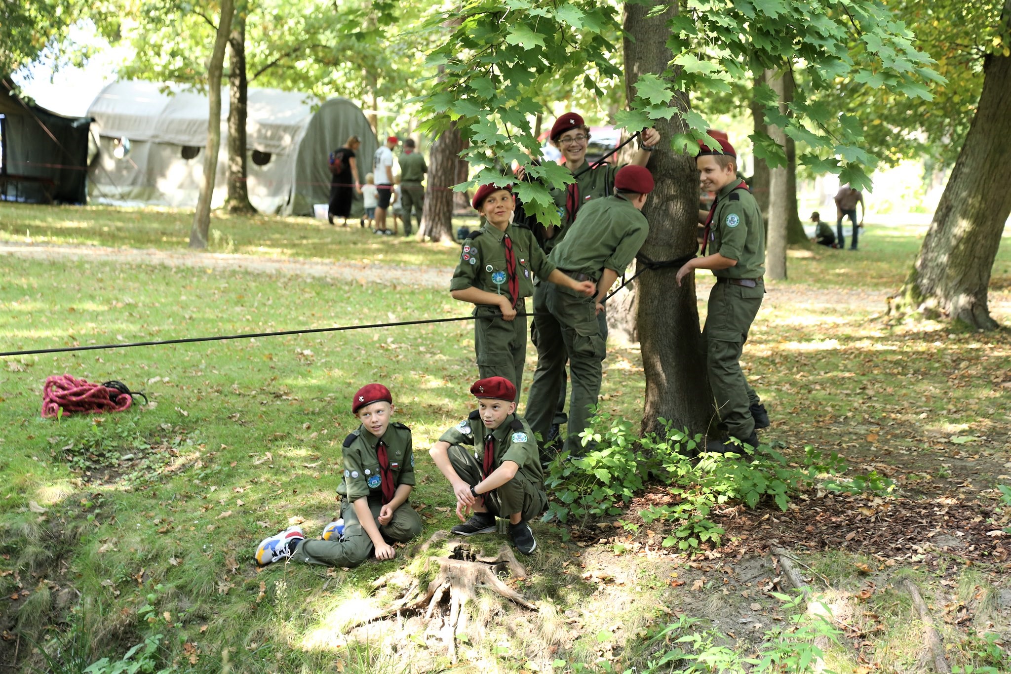 Park Zdrojowy. Grupa sześciu chłopców stoi obok siebie przy drzewie. Do drzewa przywiązana jest lina. Wszyscy są ubrani w harcerskie stroje, na głowach mają bordowe berety. 