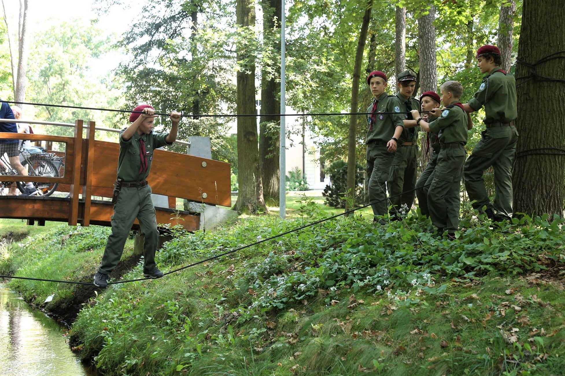 Park Zdrojowy. Chłopiec stoi na linie przywiązanej do drzewa. Obok niego na trawie stoi pięć osób. Wszyscy są ubrani w harcerskie stroje.