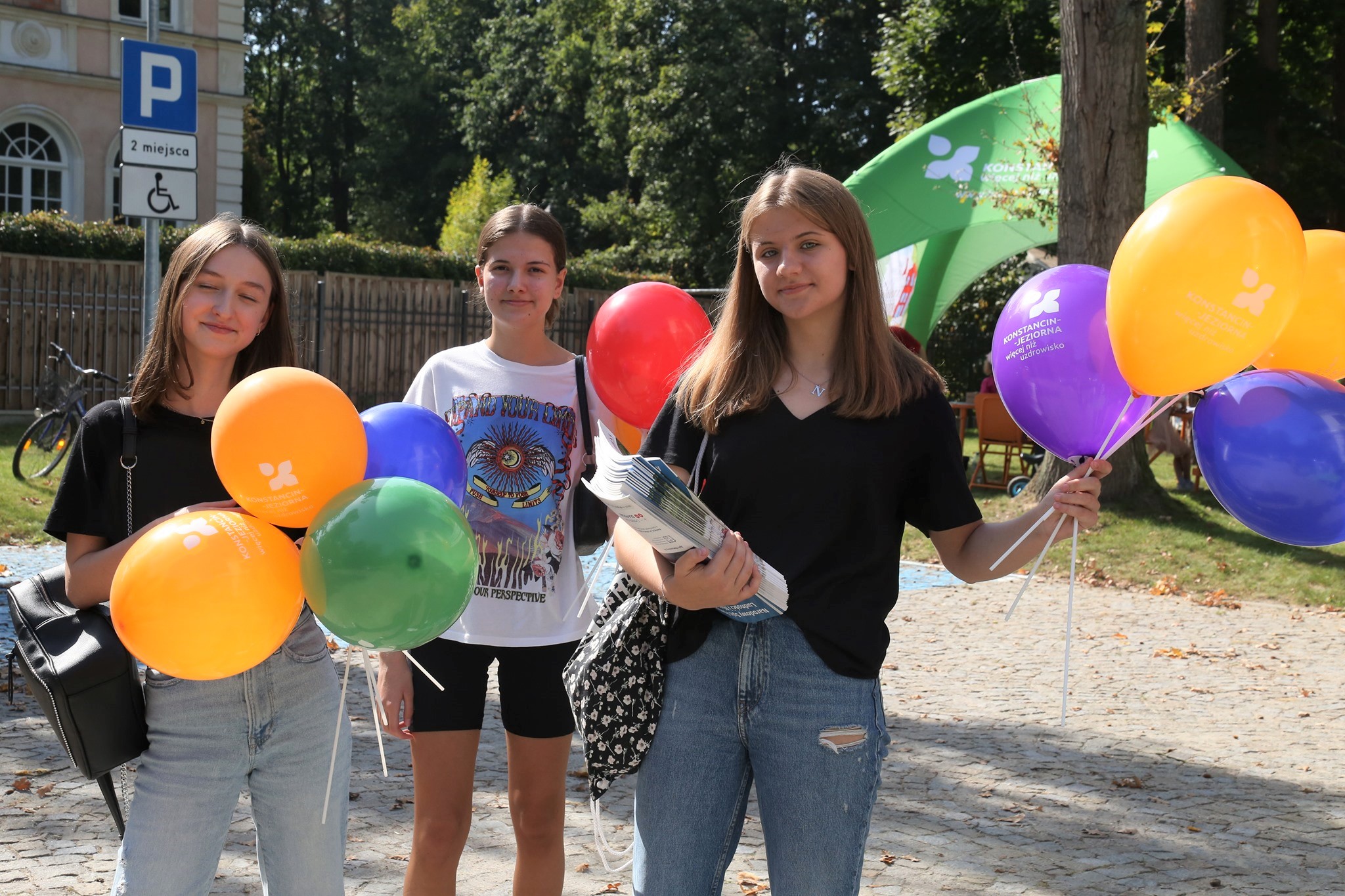 Park Zdrojowy. Trzy młode dziewczyny stoją obok siebie. Każda w rękach trzyma kolorowe balony z napisem Konstancin-Jeziorna więcej niż uzdrowisko. 
