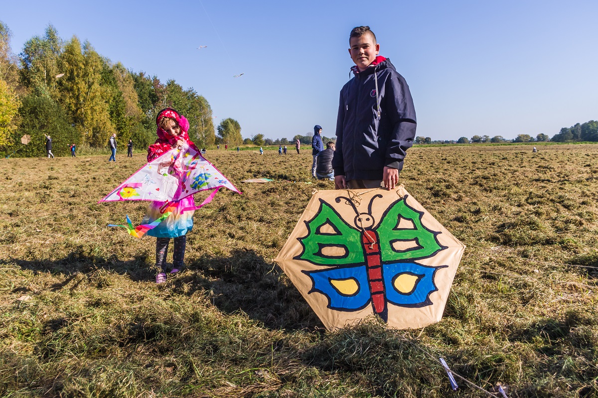 Łąki Oborskie. Chłopiec i dziewczynka w wieku szkolnym stoją obok siebie. Dziewczynka trzyma w rękach różowy latawiec w kształcie trójkąta. Chłopiec trzyma w ręku latawiec w kształcie sześciokąta , na którym namalowany jest kolorowy motyl. W tle drzewa i błękitne niebo.