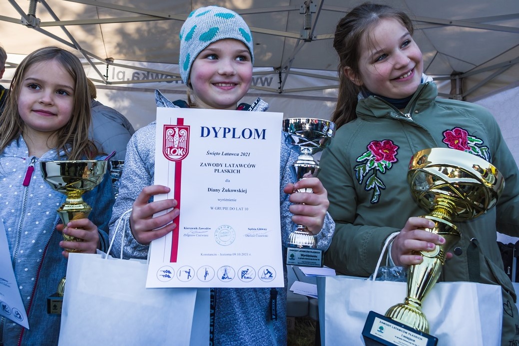 Trzy dziewczynki w wieku szkolnym stoją obok siebie w rzędzie. W rękach trzymają dyplomy i złote puchary.