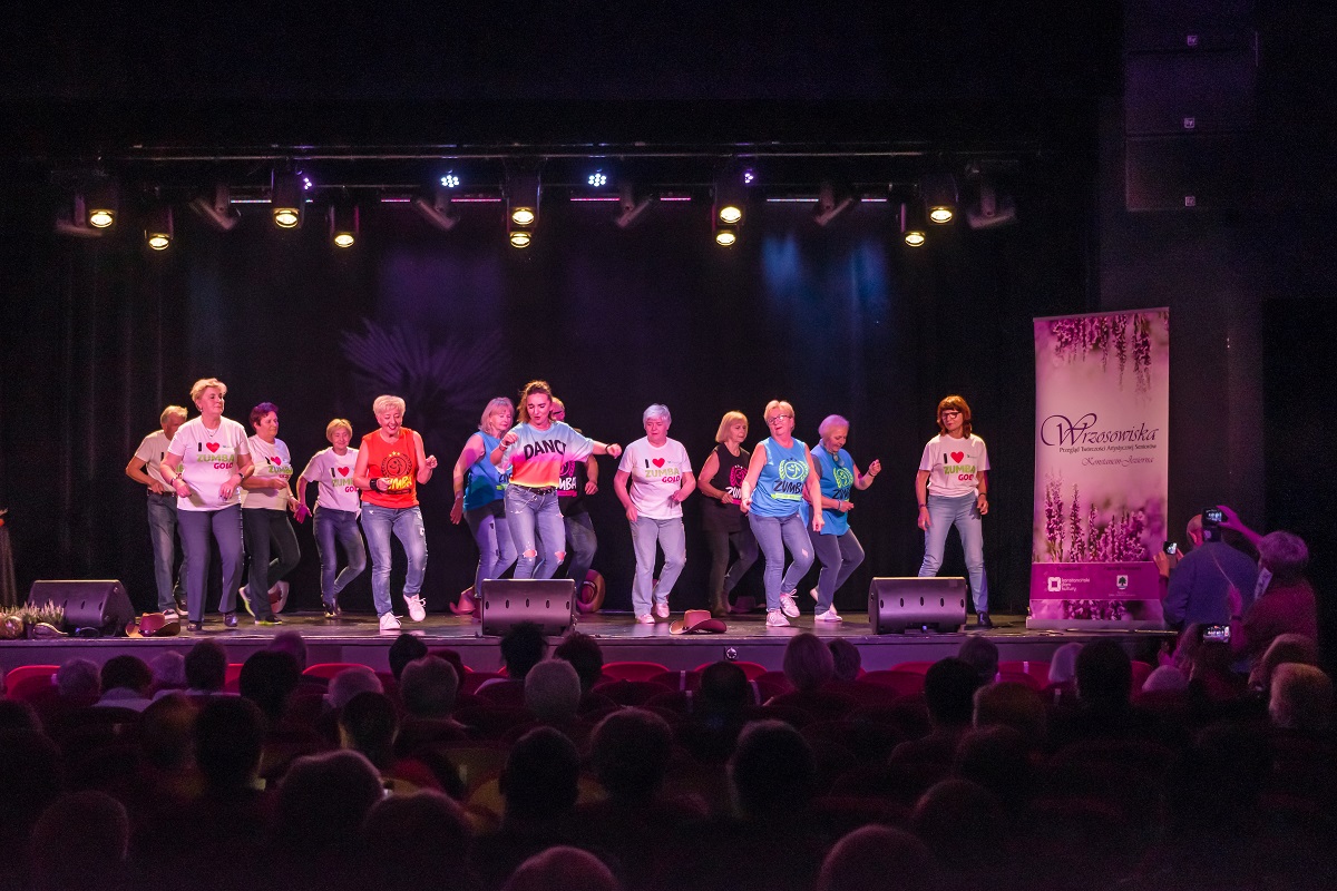 Na scenie występuje grupa taneczna. Trzynaście osób tańczy – dwanaście kobiet i jeden mężczyzna. Z przodu jest młoda kobieta, za nią osoby w średnim wieku. W tle wisi czarna zasłona. Przed nimi na fotelach siedzi publiczność.
