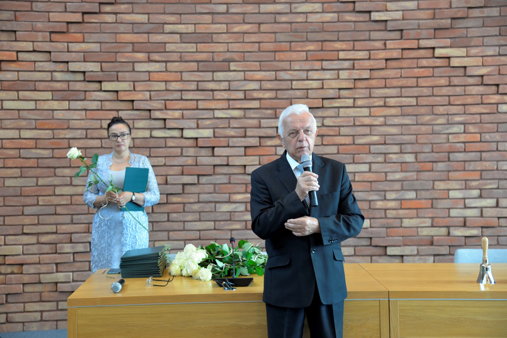 Sala konferencyjna. Mężczyzna w średnim wieku, ubrany w garnitur przemawia, w ręku trzyma mikrofon. Za nim stoi kobieta ubrana w błękitną sukienkę, w ręku trzyma białą różę. Obok na stole leżą dyplomy i bukiet białych róż.