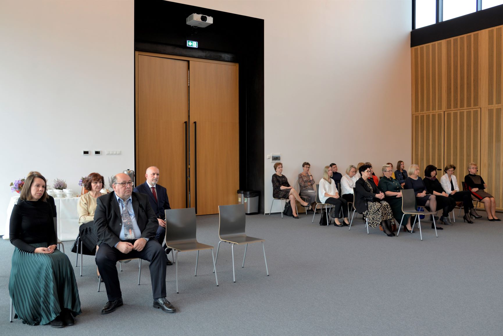 Sala konferencyjna. Około dwadzieścia osób siedzi a krzesłach po obu stronach sali. W tle są duże drewniane drzwi.