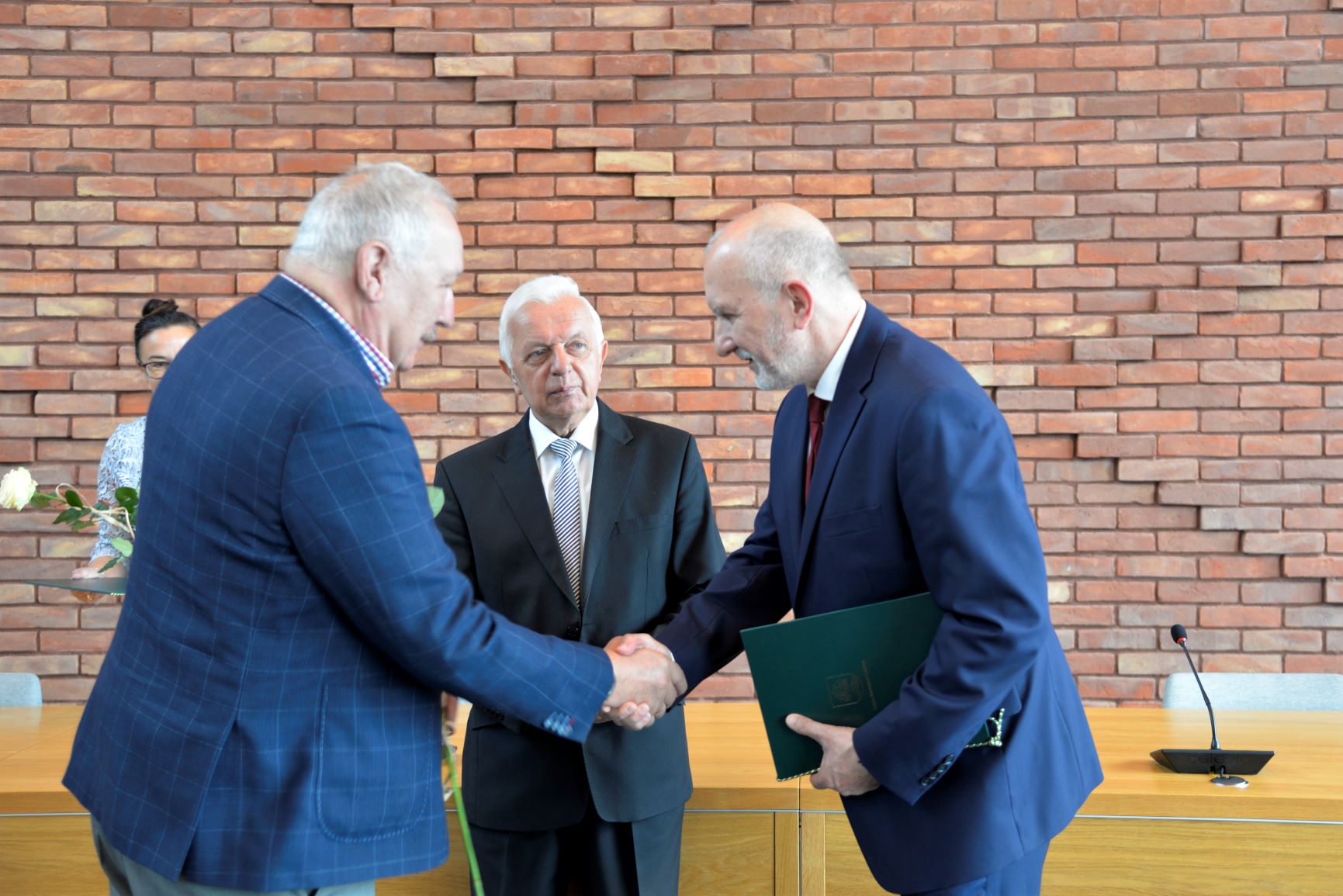 Sala konferencyjna. Mężczyzna w średnim wieku, ubrany w garnitur naprzeciwko niego stoi mężczyzna. Ściskają oni sobie prawe dłonie. Za nimi stoi mężczyzna w średnim wieku ubrany w garnitur.