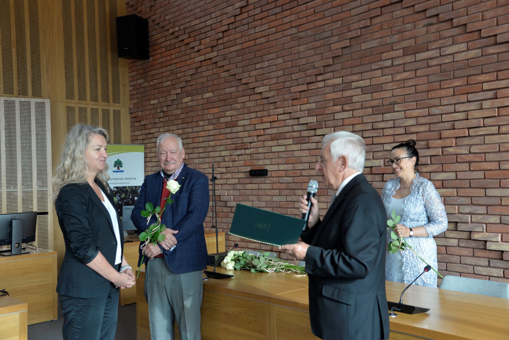 Sala konferencyjna. Mężczyzna w średnim wieku, ubrany w garnitur w jednym rękach trzyma dyplom w zielonej teczce i mikrofon. Naprzeciwko niego stoi kobieta ubrana, obok stoi mężczyzna w średnim wieku ubrany w garnitur. Za nim stoi kobieta ubrana w niebieską sukienkę.