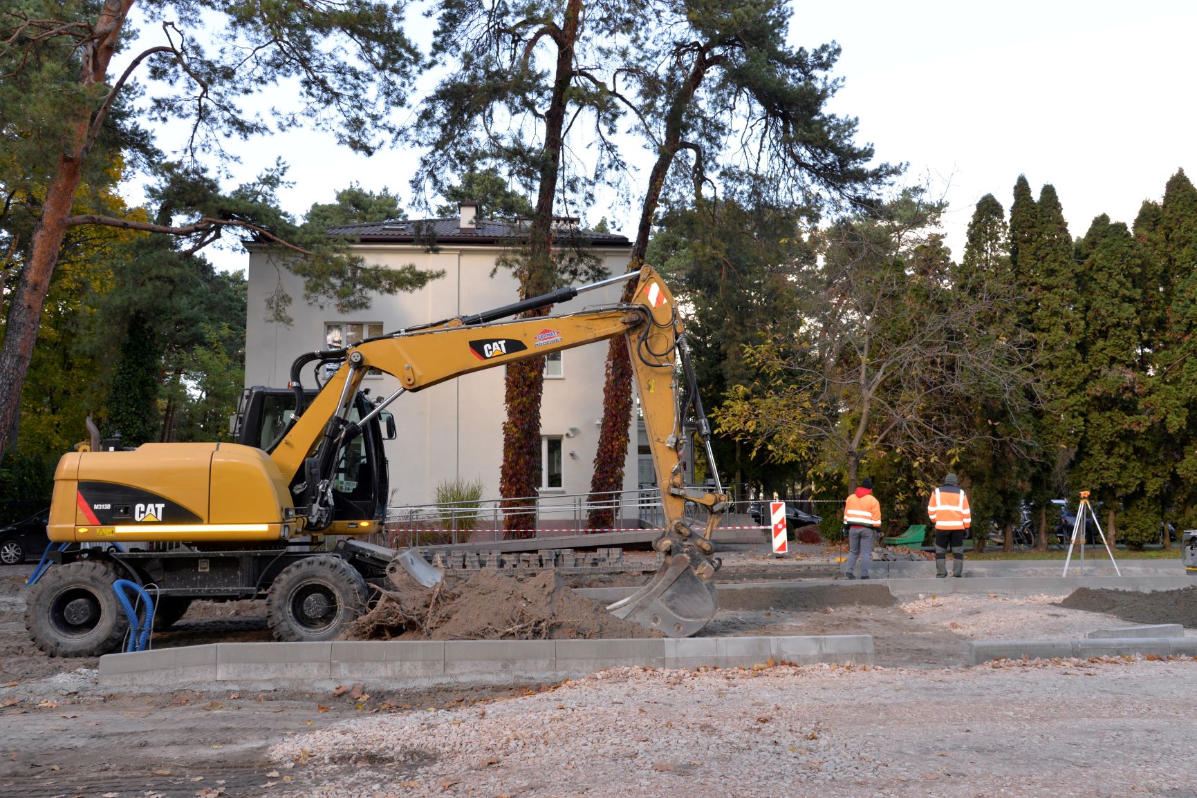Remont drogi. Widać rozkopaną ziemię, obok stoi żółta koparka. Przy koparce stoi dwóch robotników, którzy ubrani są w pomarańczowe kurtki z odblaskami.