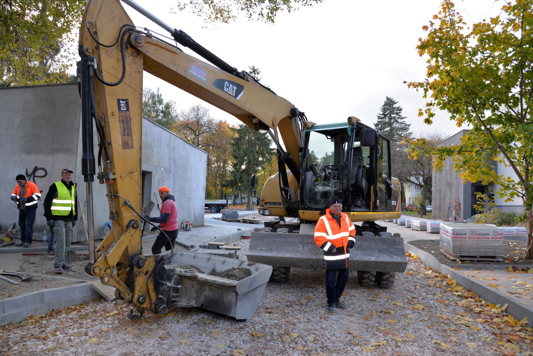 Remont drogi. Na ulicy stoi duża żółta koparka, obok niej stoi czterech robotników. Każdy ubrany jest w kurtkę z odblaskami. Wzdłuż ulicy na paletach stoi kostka brukowa owinięta folią.