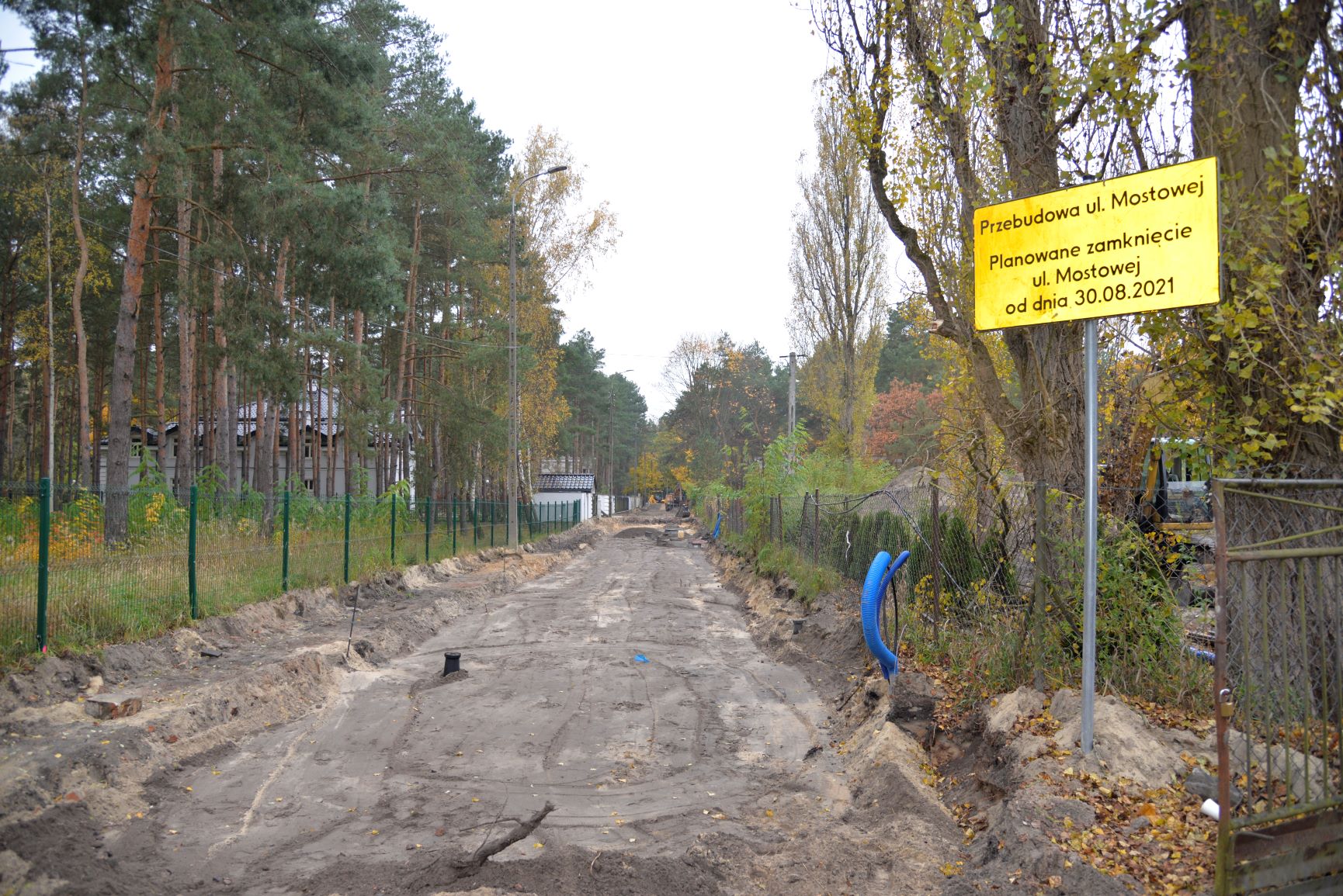 Przebudowa drogi. Widać rozkopaną ziemię. W oddali stoją dwie koparki. Po obu stronach drogi rosną drzewa. Po lewej stronie stoją latarnie. Po prawej stronie stoi znak drogowy z żółtą tabliczką, na której jest napis Przebudowa ulicy Mostowej. Planowane zamknięcie od dnia 30 sierpnia 2021.