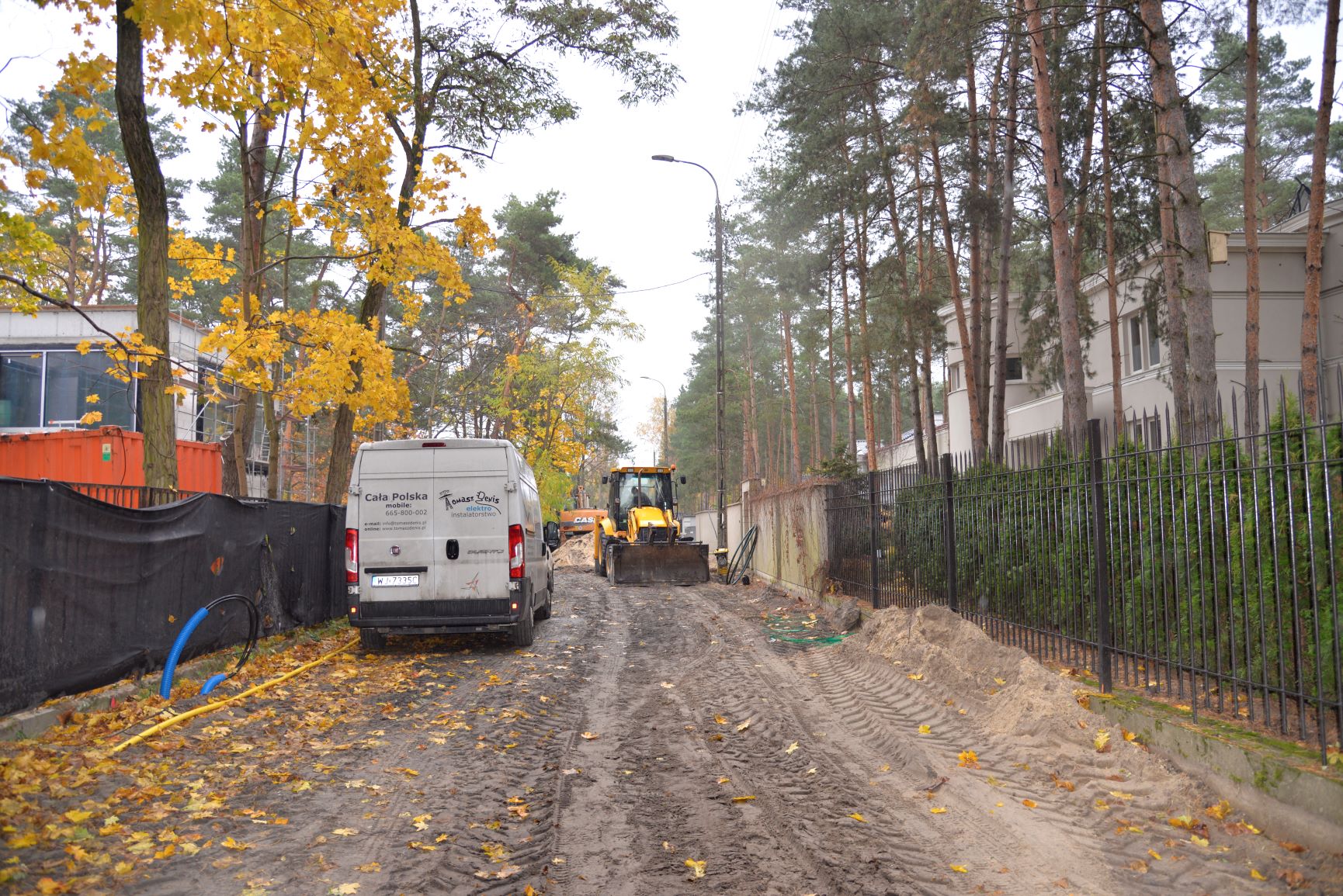 Przebudowa drogi. Widać rozkopaną ziemię, na której stoi żółta koparka. Po lewej stronie stoi biały samochód dostawczy. Po obu stronach są ogrodzenia oraz rosną wysokie drzewa.