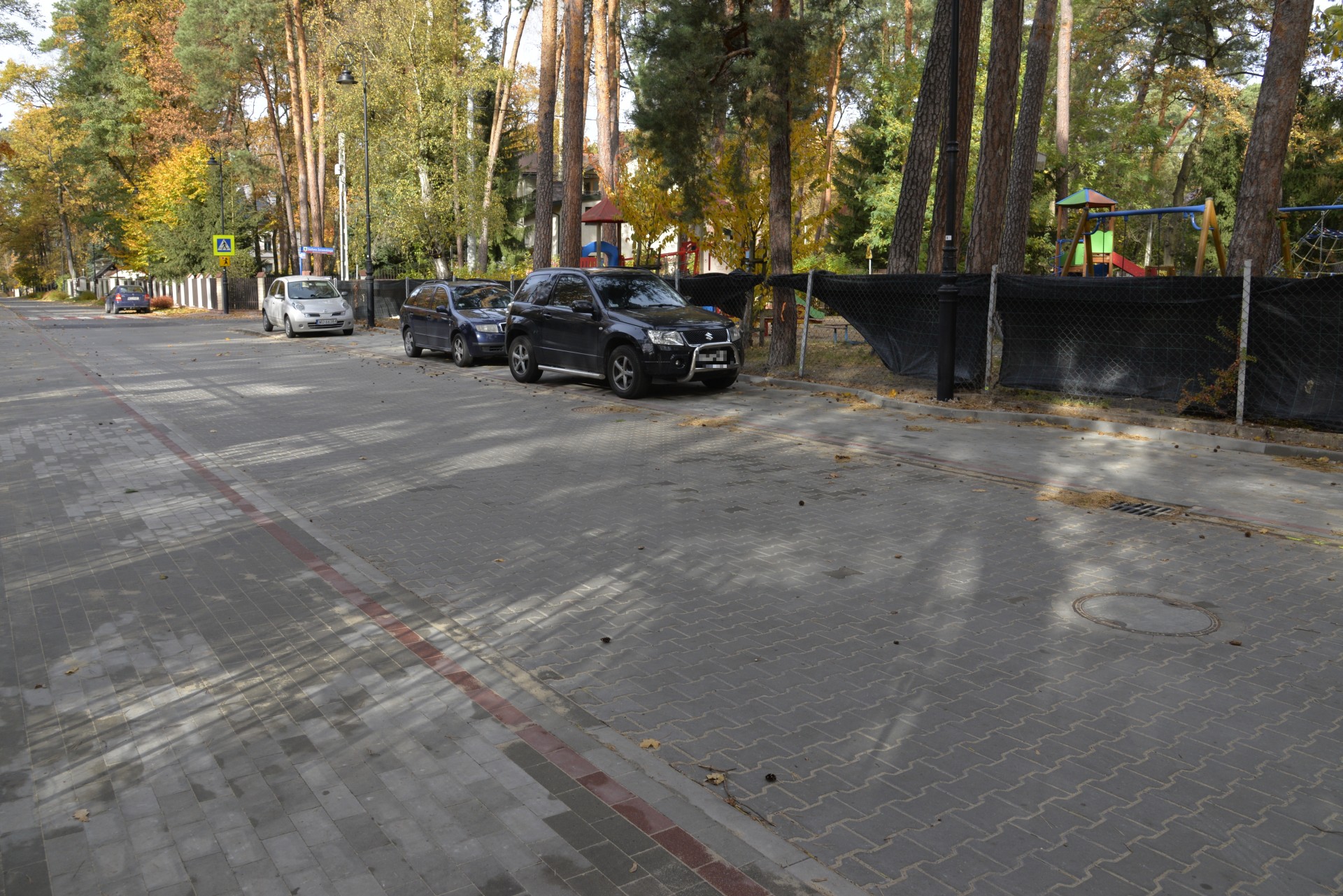 Ulica z kostki brukowej. Po prawej stronie stoją zaparkowane w rzędzie samochody. Za nimi, za ogrodzeniem, znajduje się plac zabaw. 
